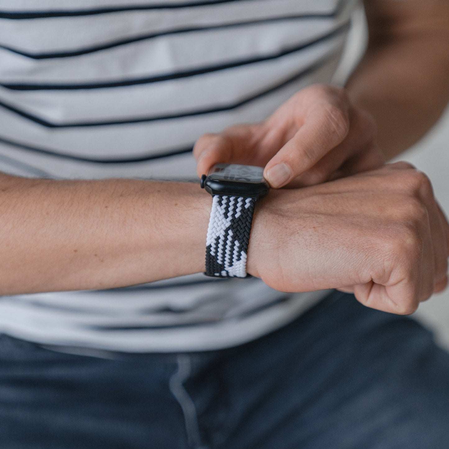 Eine Person, die ein geflochtenes Flex Loop TwoTone Armband von Arktisband für die Apple Watch trägt, berührt den Bildschirm ihrer Smartwatch. Sie trägt ein weißes Hemd mit schwarzen Streifen und blaue Jeans.
