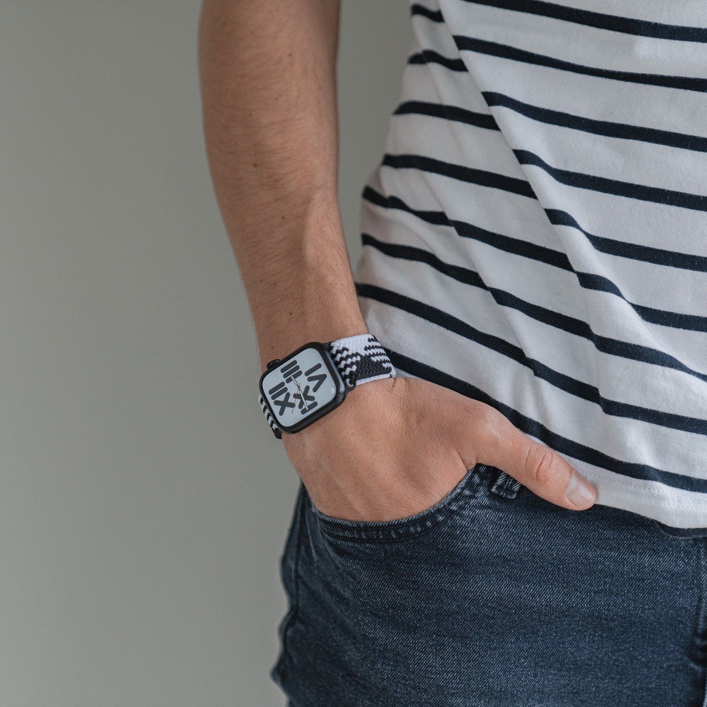 Eine Person in einem schwarz-weiß gestreiften Hemd und dunklen Jeansshorts, die rechte Hand in der Tasche, trägt außerdem das geflochtene Flex Loop TwoTone Armband der Apple Watch mit stylischem Edelstahlverschluss.