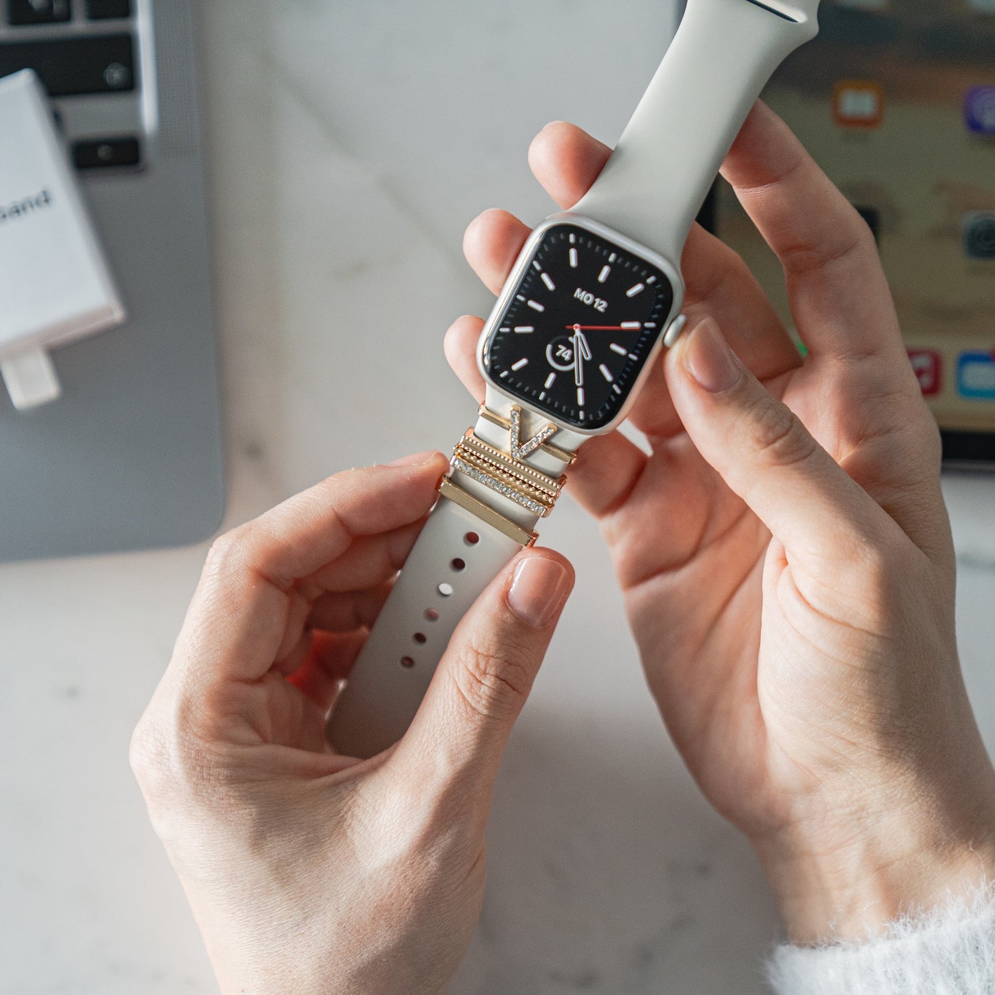 Eine Person befestigt cremefarbene arktisband Apple Watch Charms „V Style“ von arktisband an einer Smartwatch auf einem Schreibtisch für einen extravaganten Look.