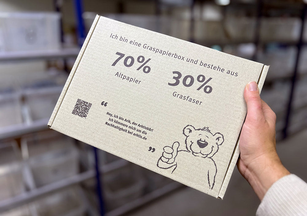 Hand hält einen Karton aus 70 % Recyclingpapier und 30 % Grasfasern mit der Illustration eines Cartoon-Bären.