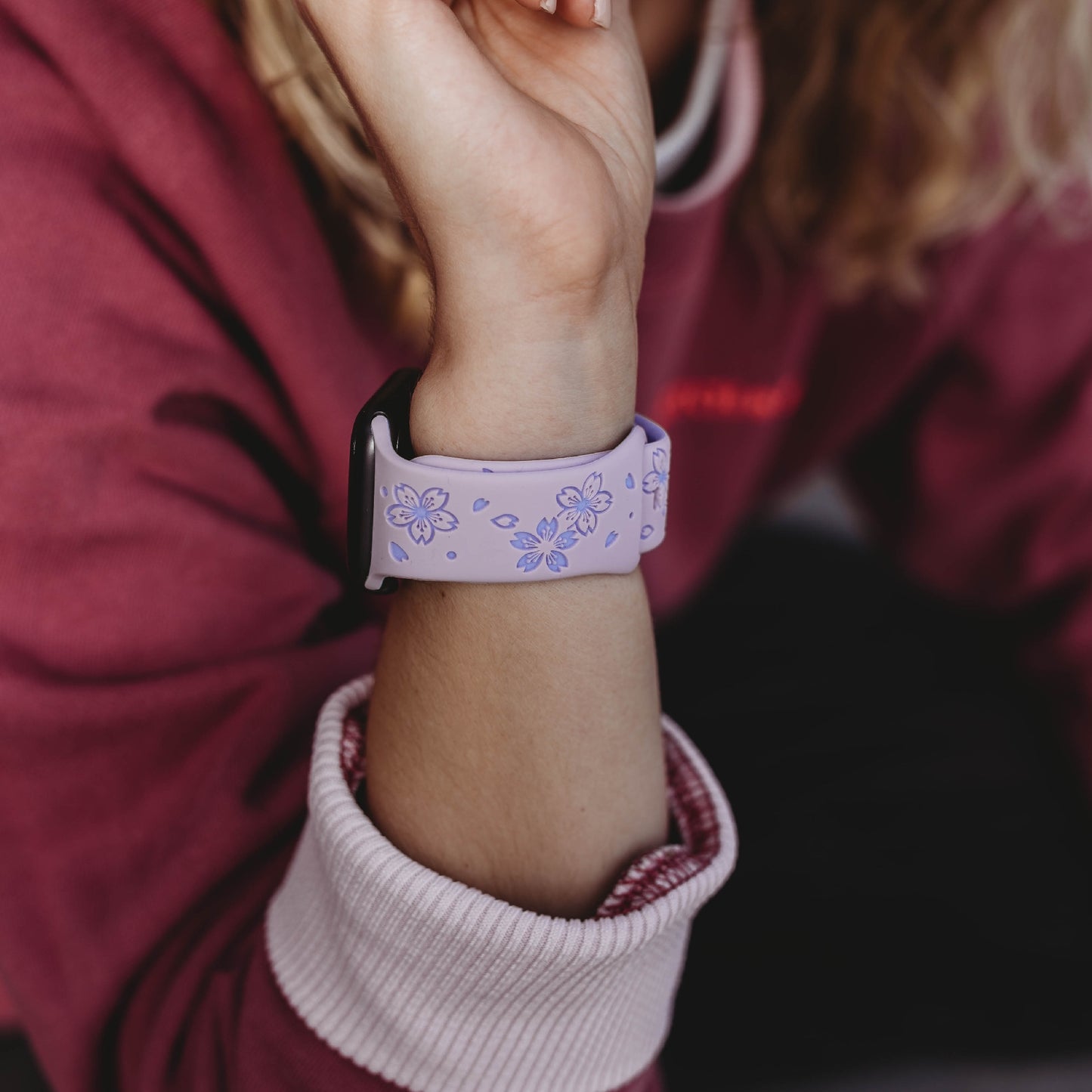 Eine Person in einem kastanienbraunen Sweatshirt mit rosa Bündchen präsentiert das arktisband Apple Watch Armband „Flores“ von arktisband in Lavendel mit blauem Blumenmuster und schweißabsorbierendem Design an ihrem Handgelenk.