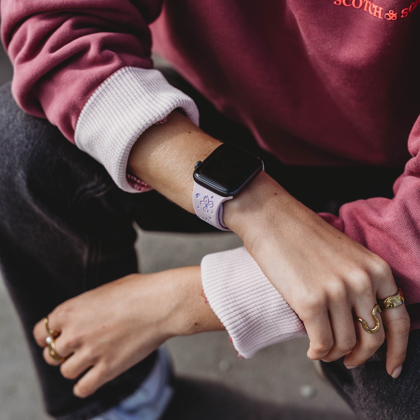 Eine Person trägt ein kastanienbraun-pinkes Sweatshirt, dunkle Jeans und goldene Ringe und stützt ihre Arme auf die Knie. An ihrem Handgelenk trägt sie ein arktisband Apple Watch Armband „Flores“ von arktisband mit einem hellvioletten Band.