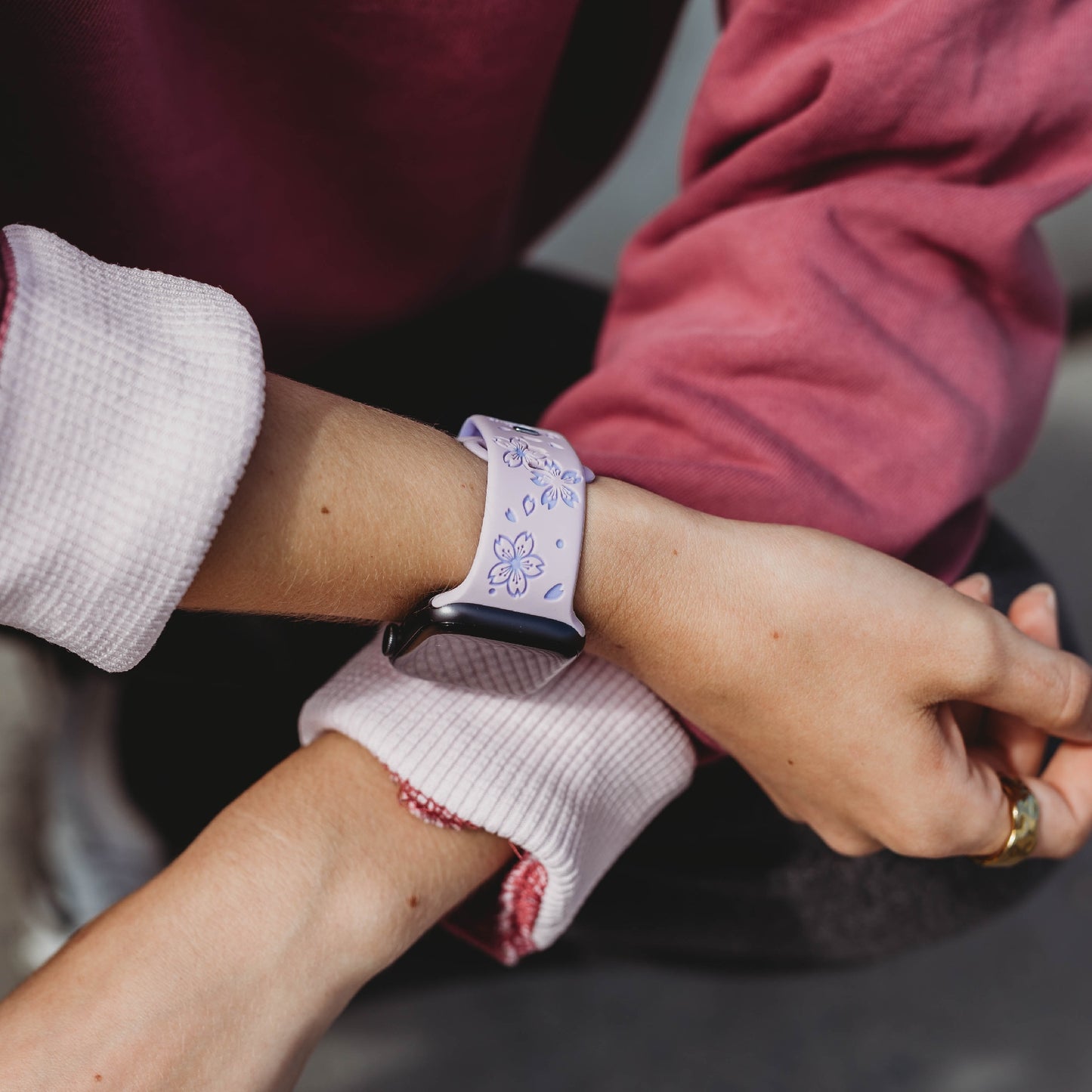 Eine Person in einem rosa-weißen Langarmshirt trägt das arktisband Apple Watch Armband „Flores“ von arktisband mit einem hellvioletten Blumenmuster am Handgelenk, wodurch das stylische, schweißabsorbierende Band hervorgehoben wird, während ihre Hände auf ihrem Schoß ruhen.