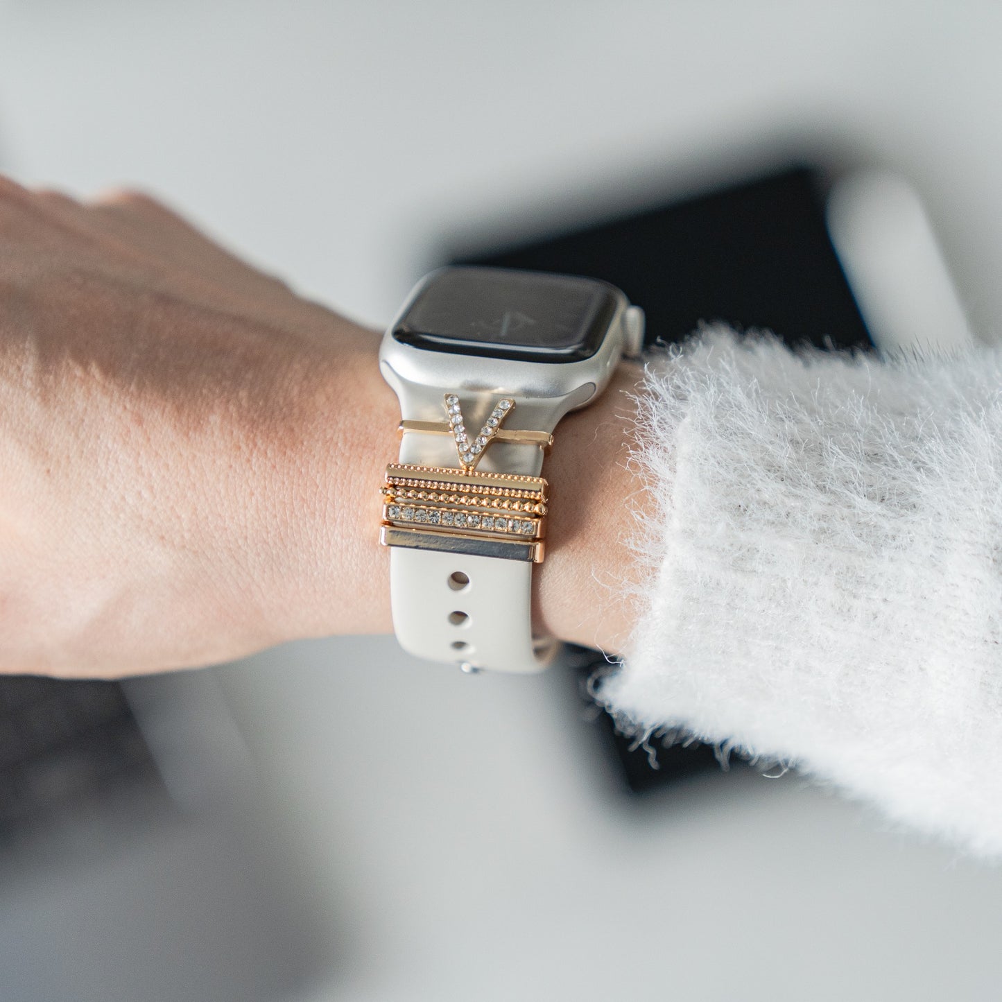Eine Person trägt eine Smartwatch mit dekorativen goldenen und silbernen Armbändern mit „V Style“-Apple-Watch-Charms von arktisband für einen extravaganten Look, mit einem verschwommenen Telefon im Hintergrund.