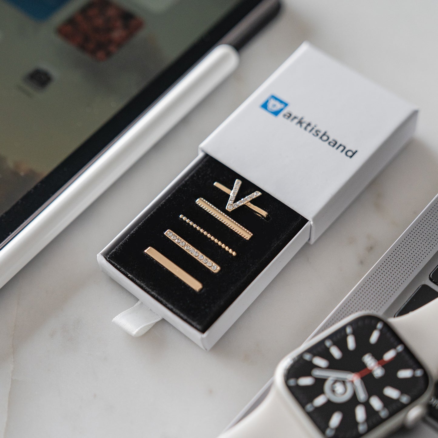 Eine Schachtel mit goldenen Ringen steht neben einer Smartwatch mit Arktisband Apple Watch Charms „V Style“, einem Laptop und einem Tablet auf dem Schreibtisch und sorgt für einen extravaganten Arbeitsplatz-Look.