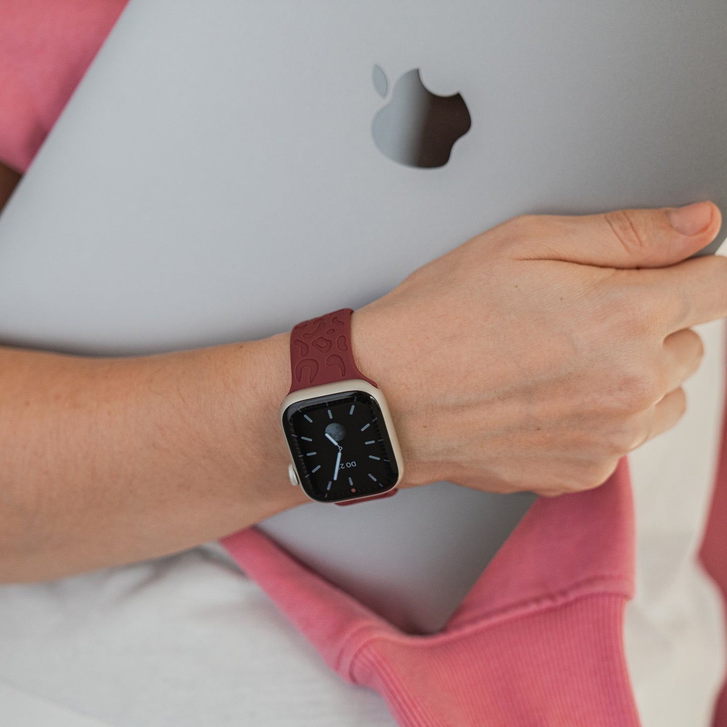 Eine Person, die ein Arktisband Apple Watch Armband „Leopard“ von Arktisband in Kastanienbraun trägt, hält einen silbernen Apple-Laptop und trägt ein rosa Sweatshirt.