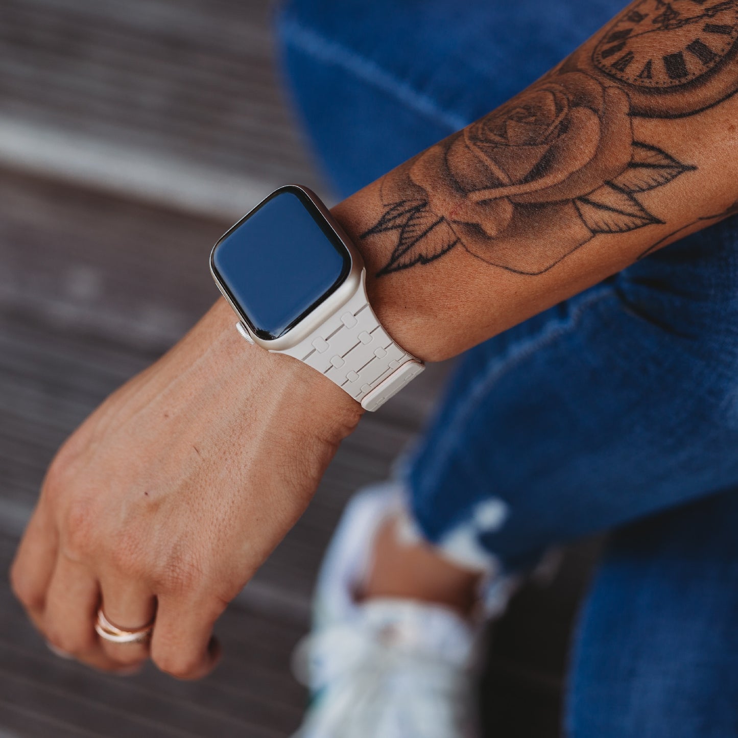 Eine Person trägt ein weißes Arktisband Apple Watch Silikonarmband "Nexus" mit integrierten Magneten und einem Ring an der linken Hand, auf dem Unterarm ist ein Rosentattoo zu sehen. Sie trägt blaue Jeans und weiße Turnschuhe.