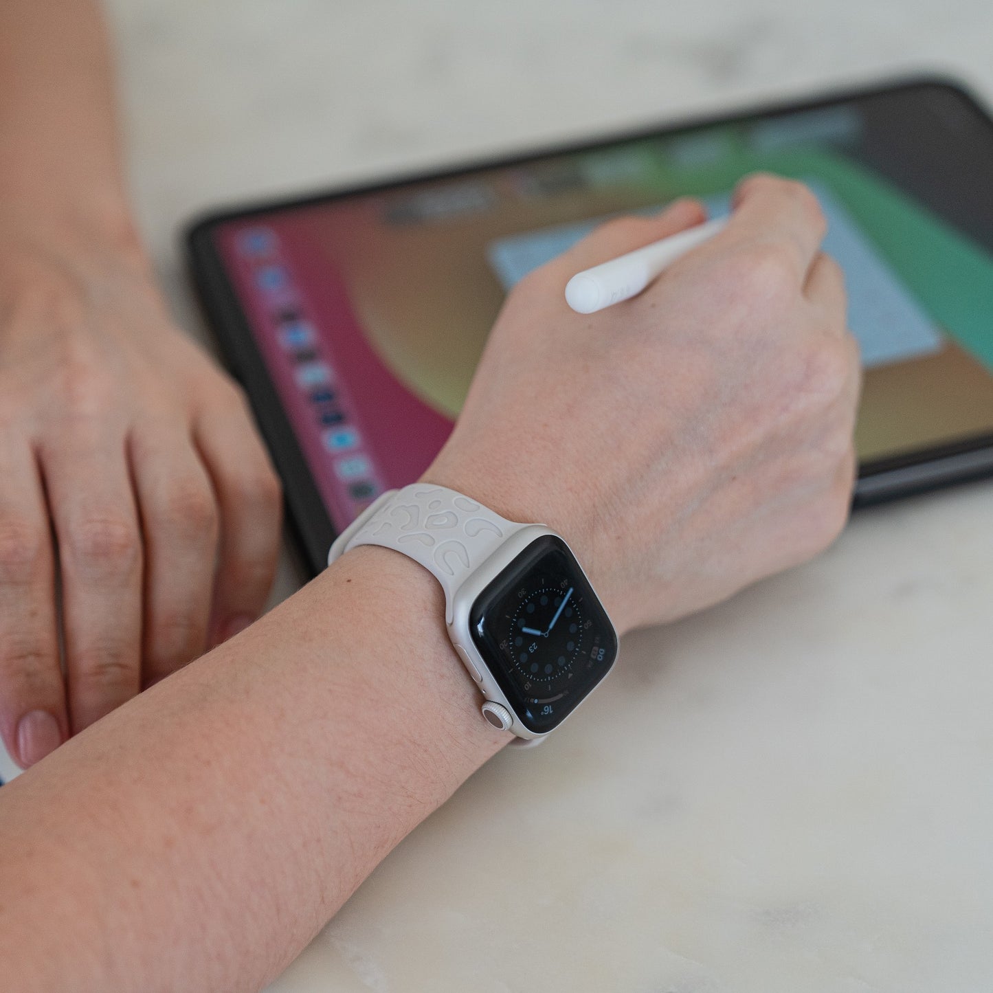 Eine Person, die ein arktisband Apple Watch Armband „Leopard“ von arktisband trägt, verwendet einen Stift zum Schreiben oder Zeichnen auf einem Tablet mit buntem Bildschirm, wobei beide Hände auf einer hellen Oberfläche ruhen.