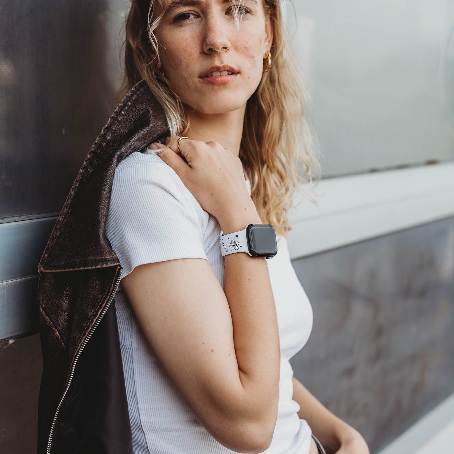 Eine Person mit langen, welligen blonden Haaren, einem weißen T-Shirt und einer dunklen Jacke über der Schulter steht draußen. An ihrem linken Handgelenk trägt sie ein arktisband Apple Watch Armband „Flores“ von arktisband und blickt in die Kamera.