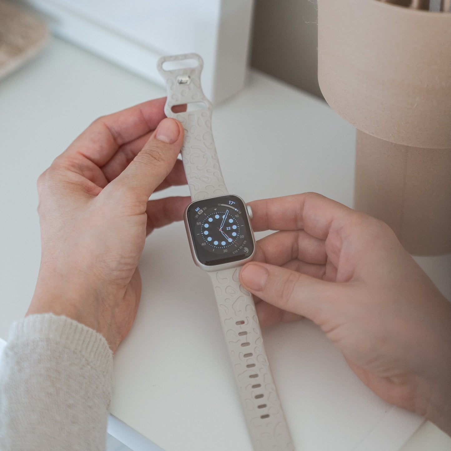Eine Person hält ein Arktisband Apple Watch Armband „Leopard“ – ein weißes Silikonarmband mit Leopardenmuster für die Apple Watch. Die Uhr hat ein analoges Zifferblatt. Die Person befindet sich in einem Raum in der Nähe einer weißen Fläche.