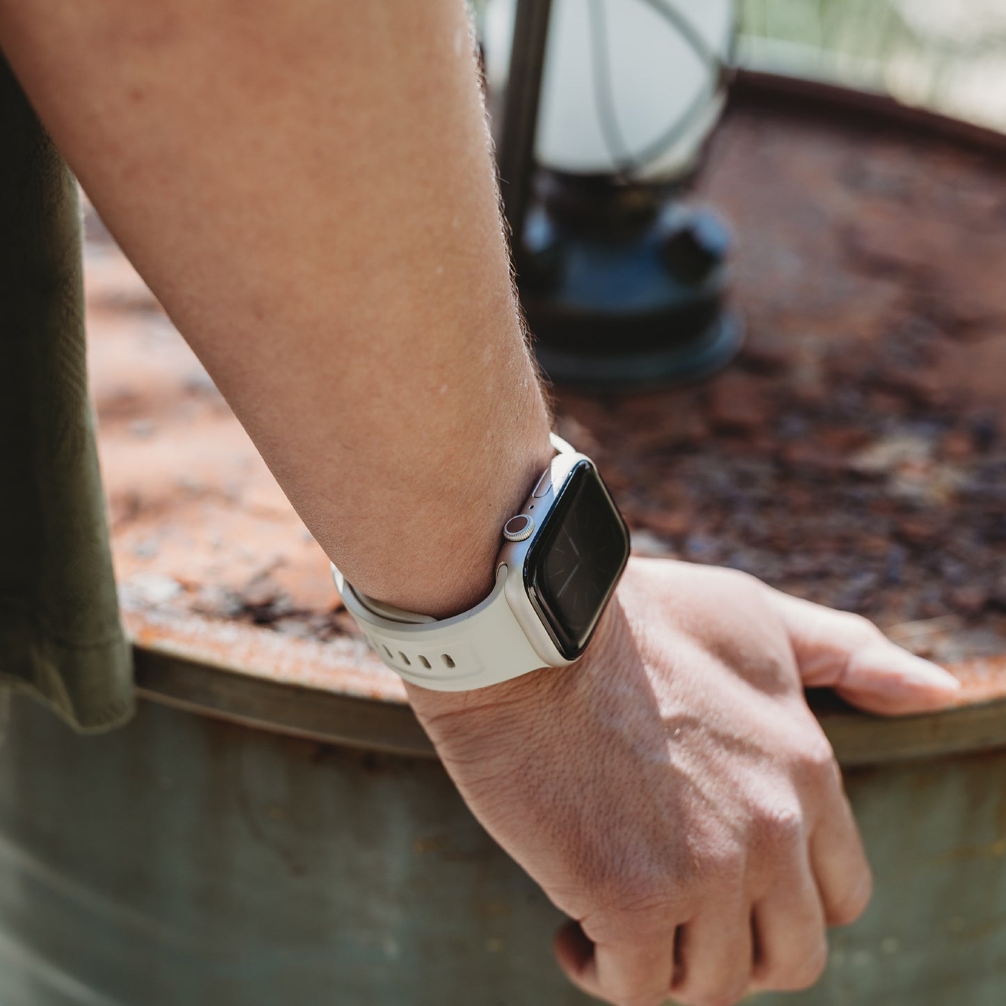 Eine Person, die das Arktisband Apple Watch FKM Sportarmband „Sweat“ trägt, legt ihre Hand auf ein verrostetes Metallfass. Das weiße Arktisband sticht hervor, während im Hintergrund eine Laterne auf dem Fass sitzt.