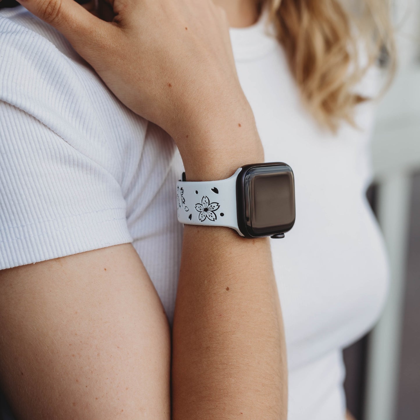 Eine Person in einem weißen Kurzarmhemd legt ihren Arm auf die Schulter und zeigt ein arktisband Apple Watch Armband „Flores“ mit Blumenmuster. Der Bildschirm der Uhr ist ausgeschaltet.