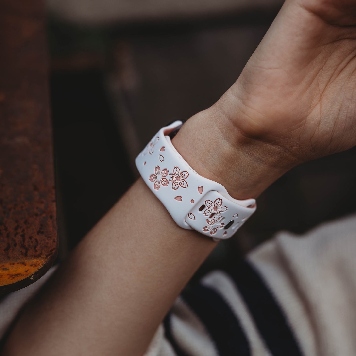 Das Handgelenk einer Person ist mit einem Arktisband Apple Watch Armband „Flores“ mit rosa Blumenmuster geschmückt. Der Arm ruht auf einer Holzoberfläche und die Person trägt einen cremefarbenen Ärmel mit einem dunklen Streifen.