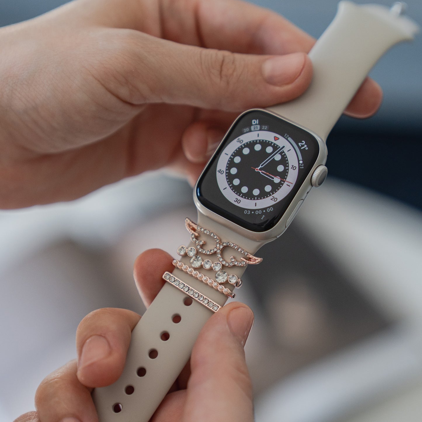 Eine Person passt ihr Arktisband mit „Diamond Wings“-Apple-Watch-Charms in Roségold an, die mit Juwelen besetzte Akzente auf einem weißen Band aufweisen, während die Smartwatch ein analoges Zifferblatt in Schwarzweiß anzeigt.