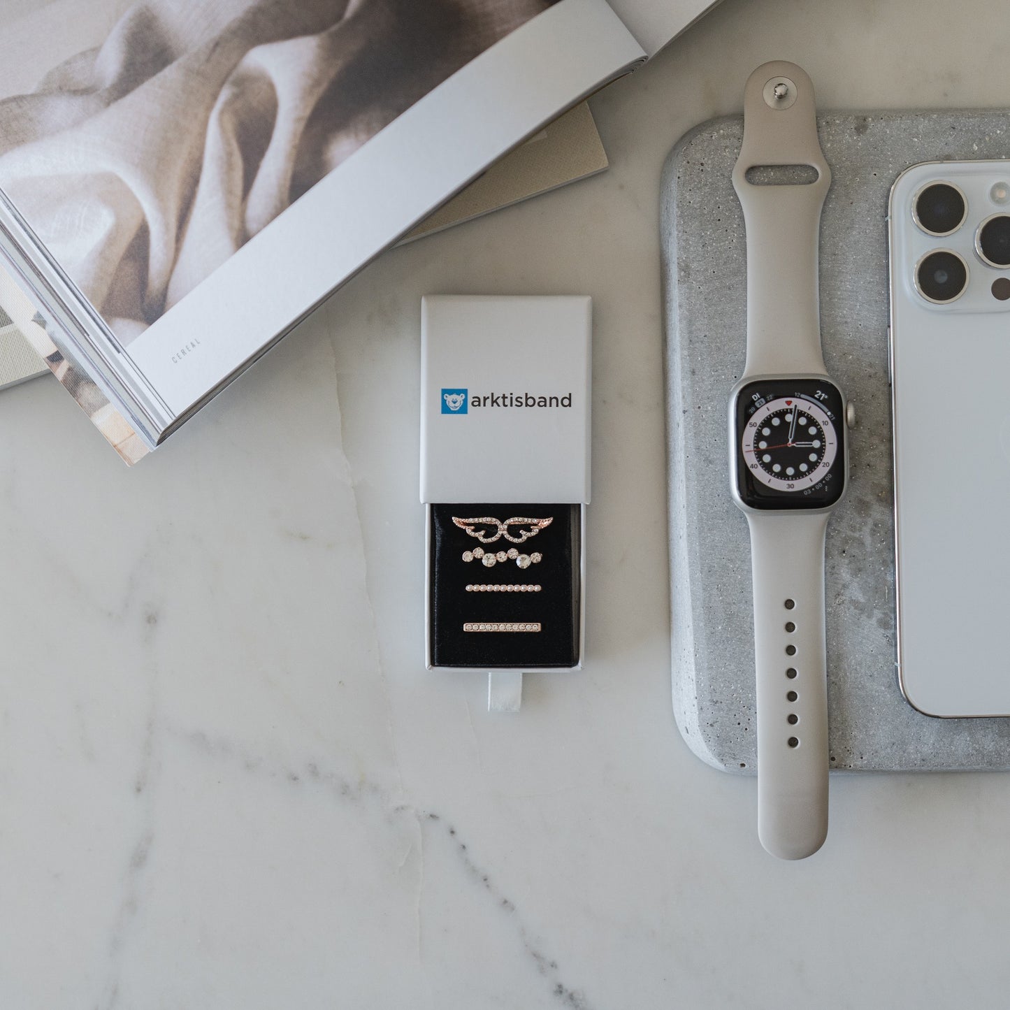 Eine hellgraue Smartwatch mit schwarzem Bildschirm liegt neben einem Telefon, einem grauen Tablett, einer Broschüre und einer offenen Arktisband-Box mit kleinen Uhrenaccessoires und Arktisband-Apple-Watch-Charms „Diamond Wings“ in Roségold auf einer weißen Marmoroberfläche.