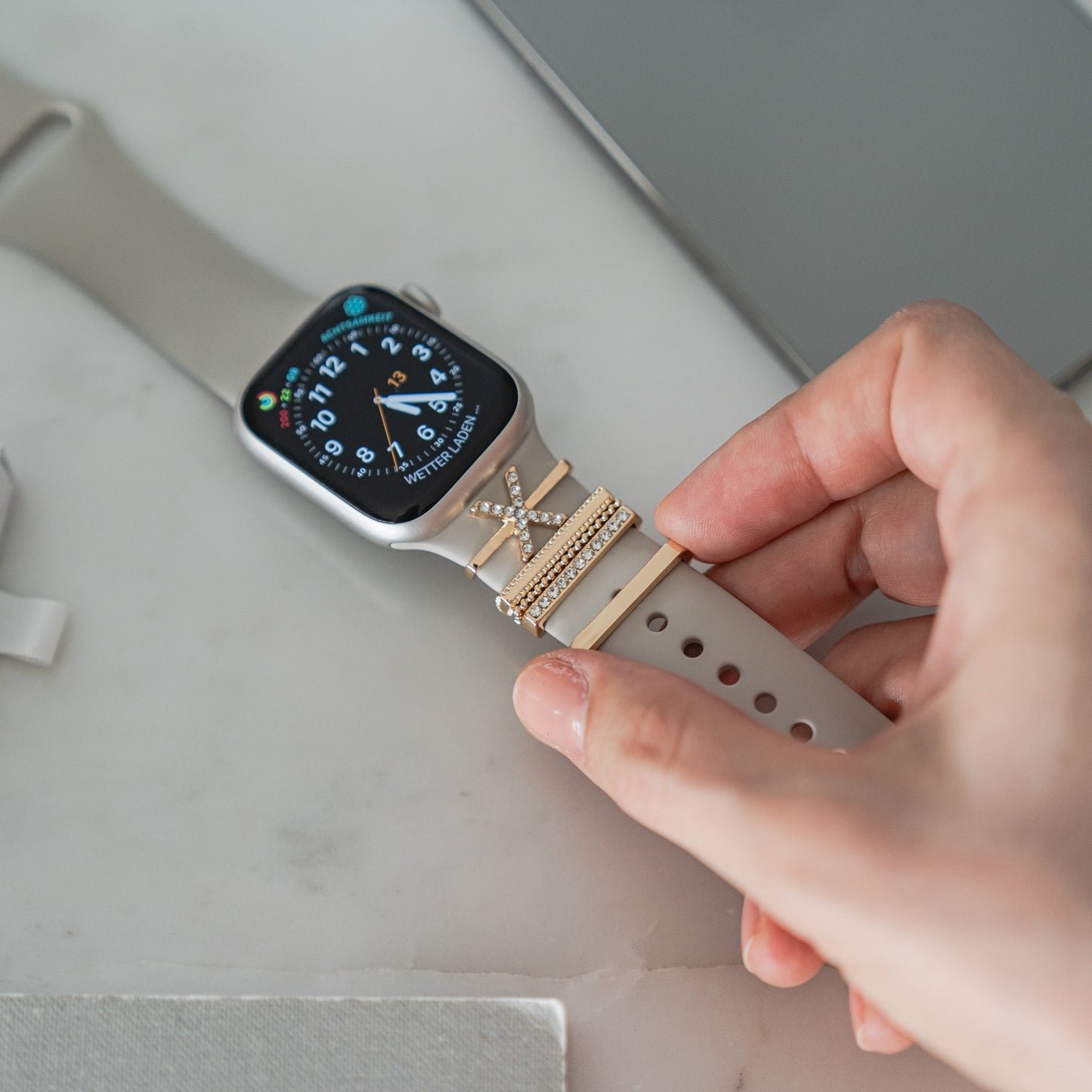 Eine Hand passt ein beiges Smartwatch-Armband an, das mit den Apple Watch Charms „X Style“ von Arktisband verziert ist – ein schickes Accessoire von Arktisband, das jeden Look aufwertet.