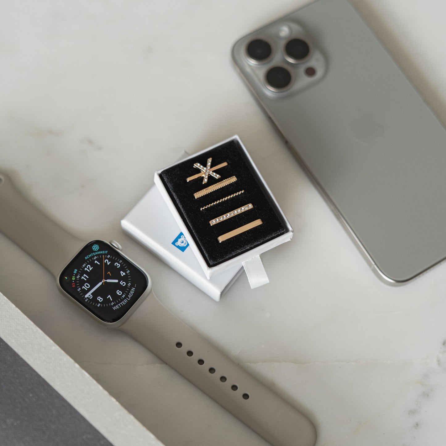 Eine Apple Watch mit arktisband „X Style“-Charms, goldener Schmuck in einer Box und ein silbernes Smartphone liegen auf einer weißen Oberfläche und präsentieren arktisband als die perfekte stylische Accessoire-Kollektion.