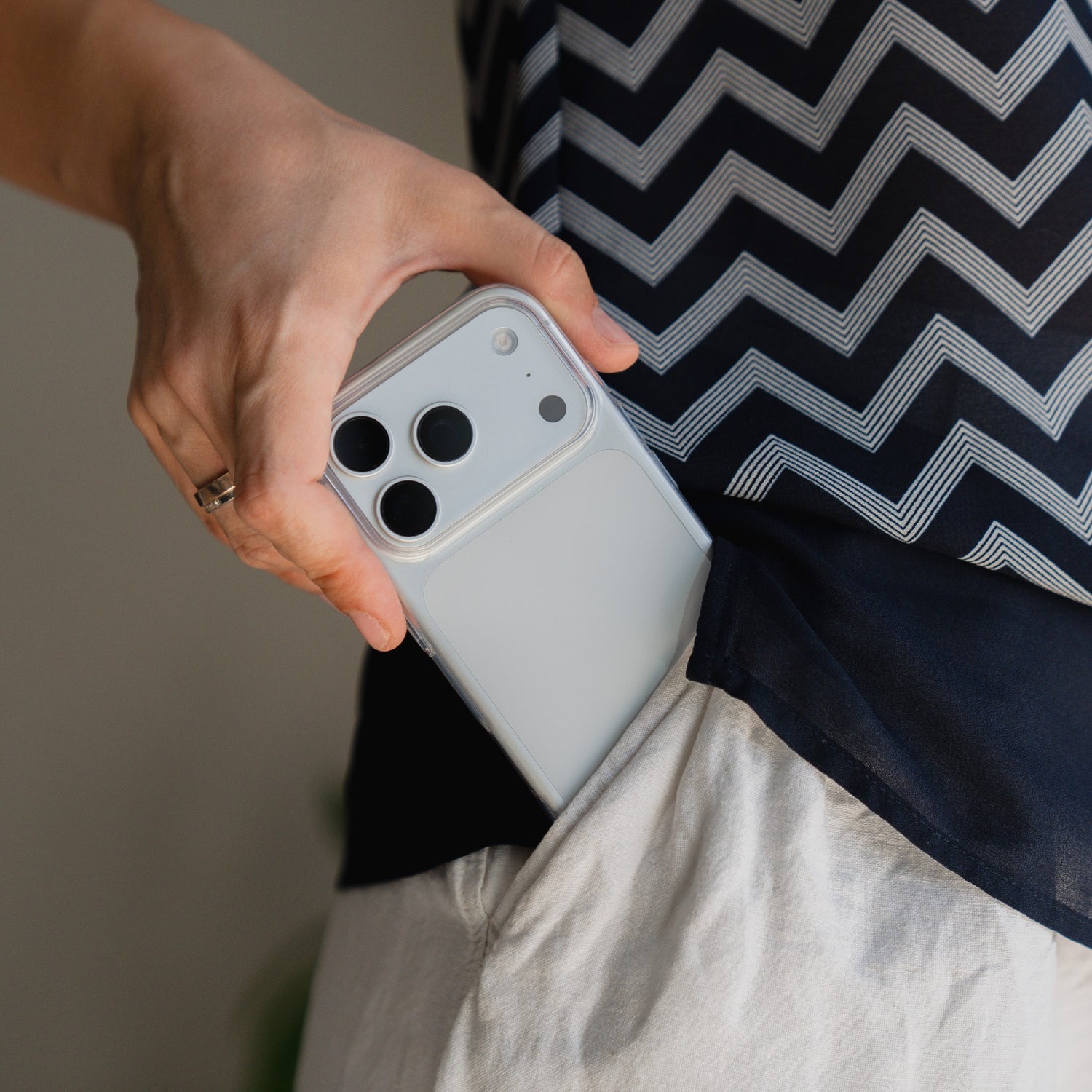Eine Hand steckt ein weißes Smartphone, das durch das arktis iPhone 17 Pro Max ZERO Case geschützt ist, in die Tasche einer hellen Hose, wobei ein Hemd mit Zickzack-Muster zu sehen ist.