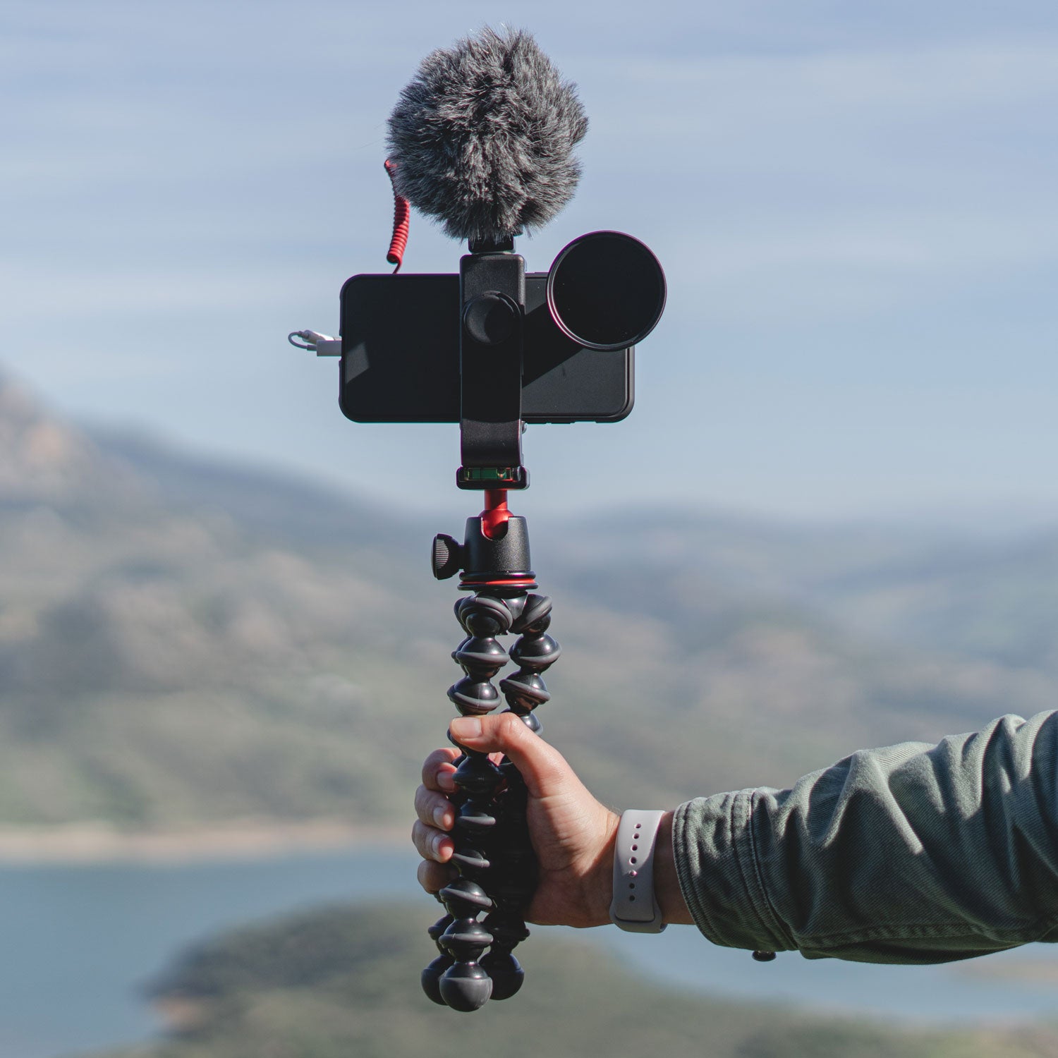 Eine Hand hält ein Smartphone auf einem flexiblen Stativ mit einem Wolffilms CPL-Polfilter und Mikrofon, im Freien in der Nähe von Bergen und Wasser.