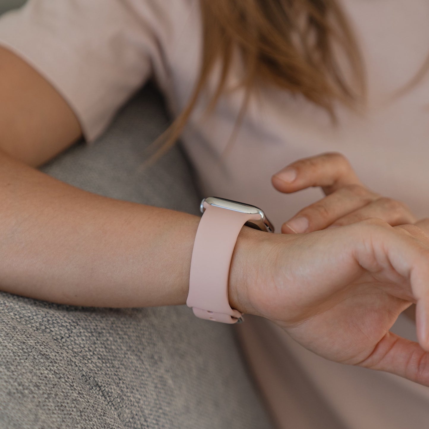 Eine Person in einem hellrosa Kurzarmhemd sitzt auf einem grauen Sofa und berührt ihre Smartwatch mit einem arktisband Apple Watch Silikonarmband „Aqua“ von arktisband am Handgelenk.