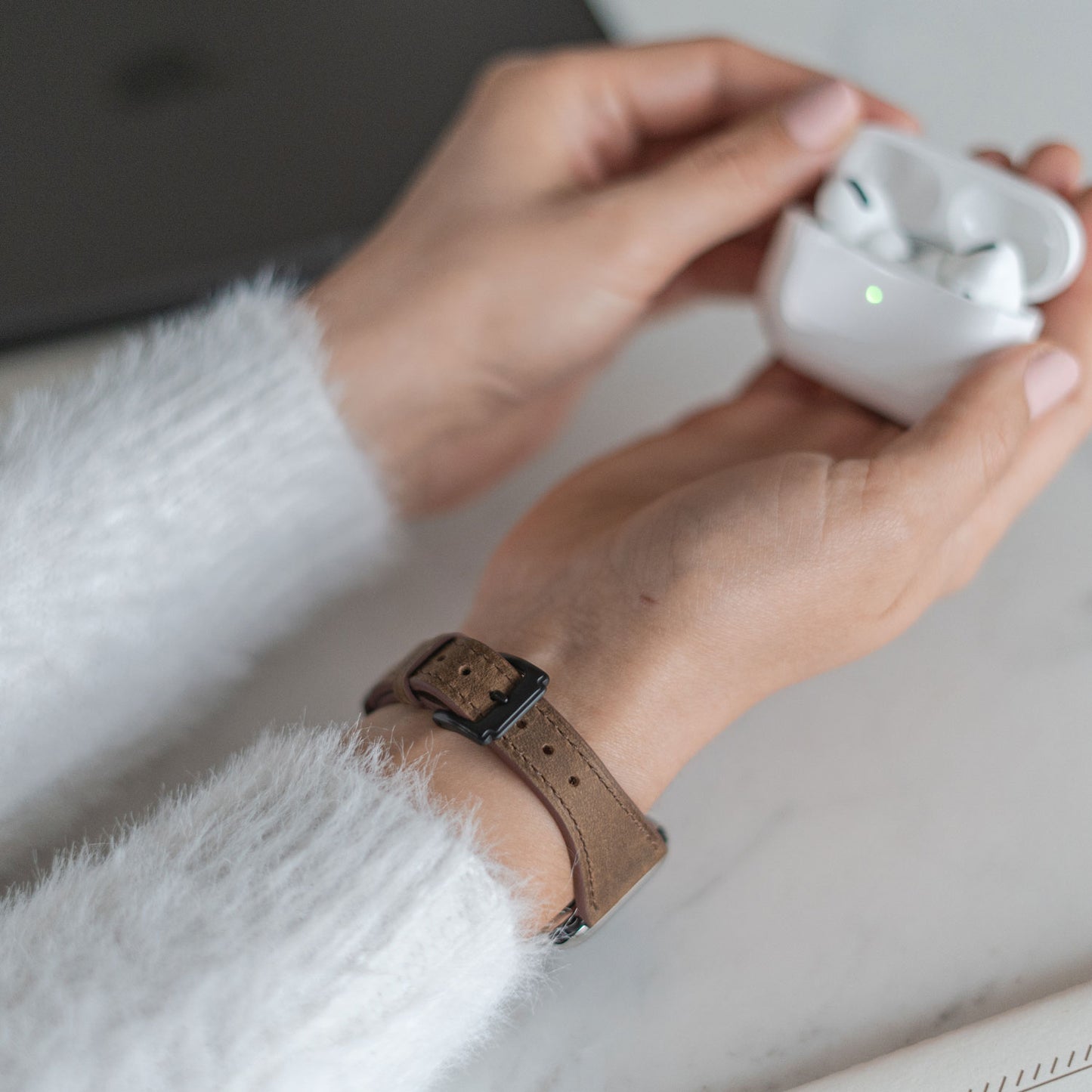 Eine Person, die das arktisband Apple Watch Lederarmband „Napoli“ von arktisband trägt, öffnet ein weißes kabelloses Ladeetui für Ohrhörer auf einem Tisch, daneben ein graues Gerät und ein Teil eines Notebooks für einen minimalistischen Look.