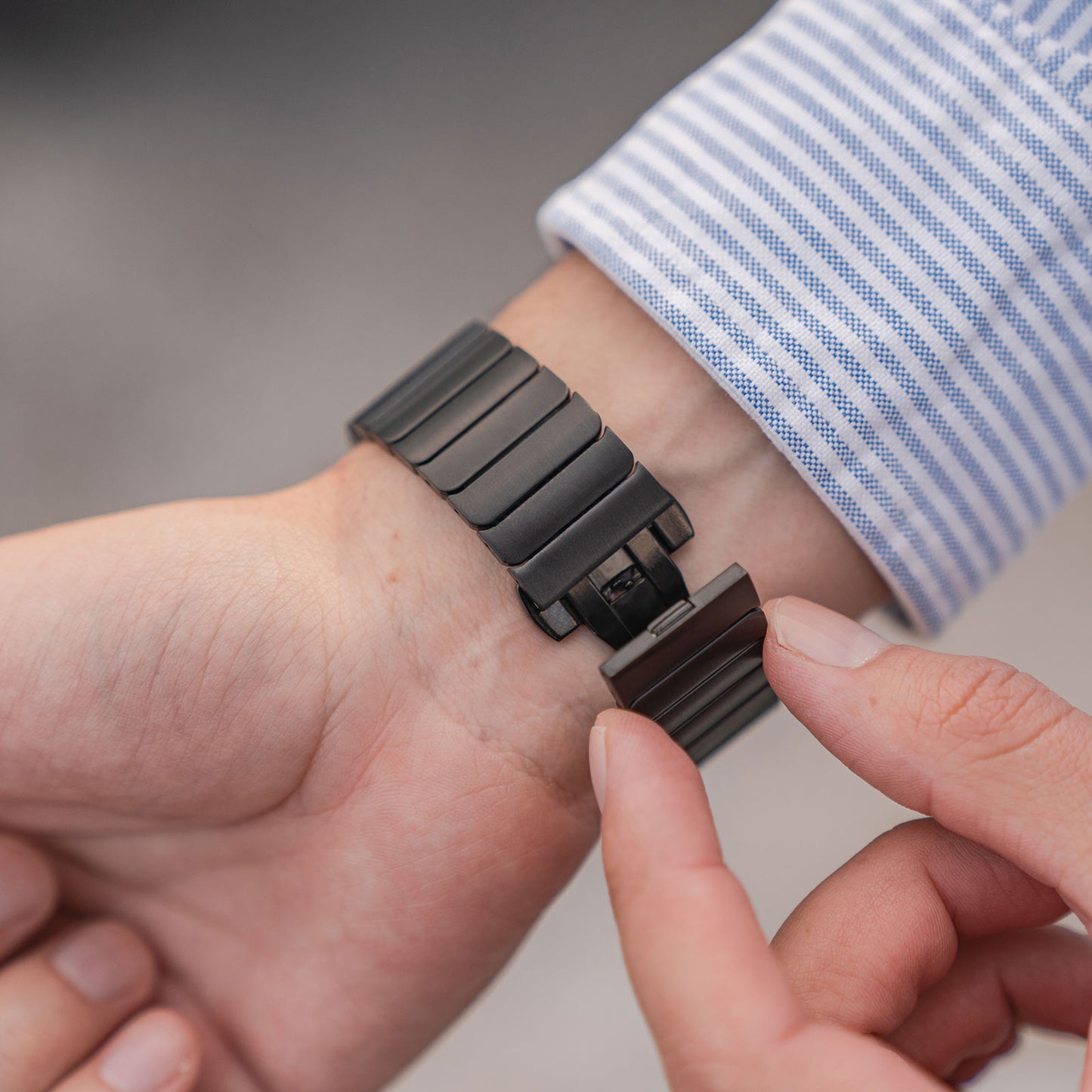Eine Person in einem blau-weiß gestreiften Hemd befestigt mit beiden Händen das arktisband Apple Watch Edelstahl Gliederarmband „Iconic“ von arktisband um ihr Handgelenk.