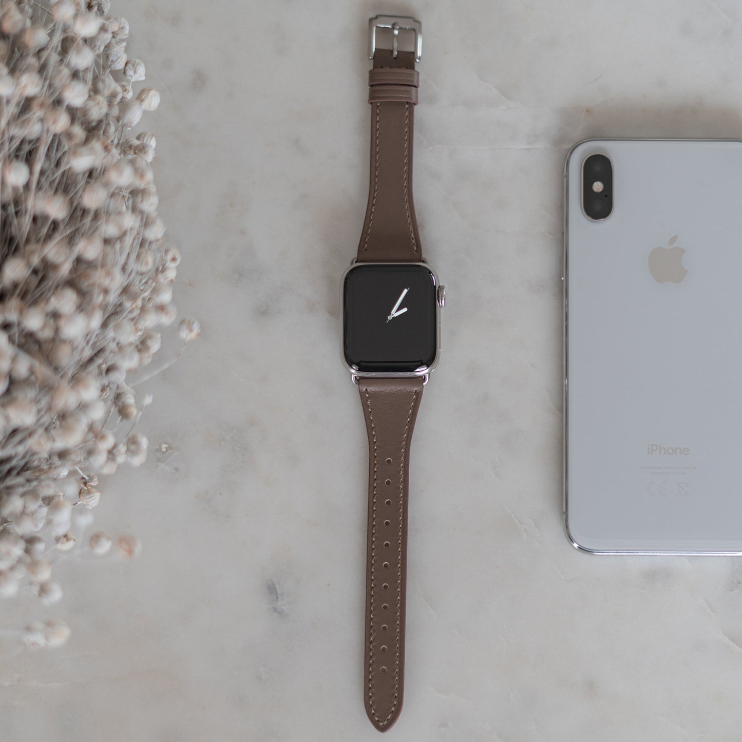 Eine arktisband Apple Watch mit dem Lederarmband „Napoli“ von arktisband liegt im minimalistischen Stil flach auf Marmor neben einem silbernen iPhone, links ist teils ein Bund getrockneter Blumen zu sehen.