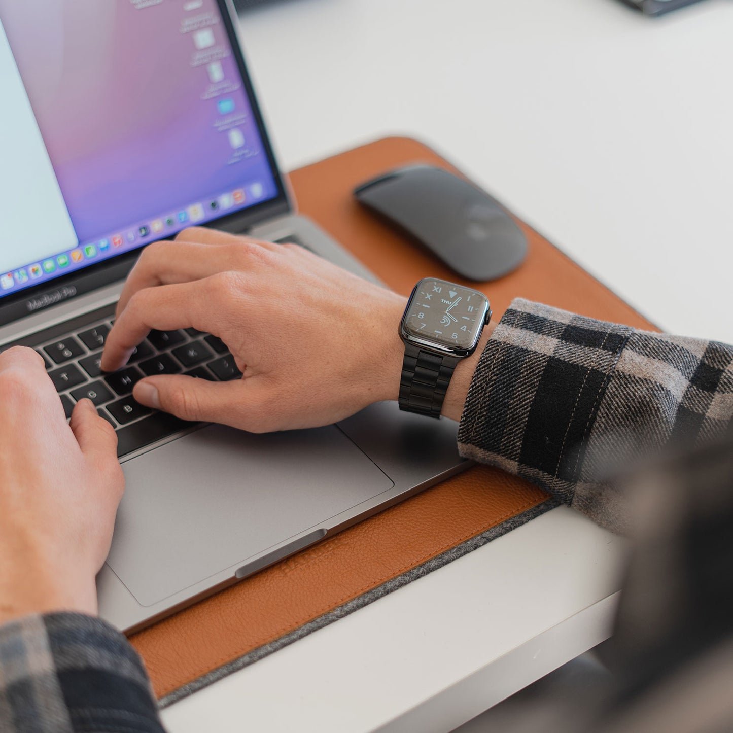 Eine Person in einem schwarz-weiß karierten Hemd tippt auf einem MacBook, daneben steht eine schwarze kabellose Maus. Sie trägt das Arktisband Apple Watch Edelstahl Gliederarmband „Edition S“ im minimalistischen Design.
