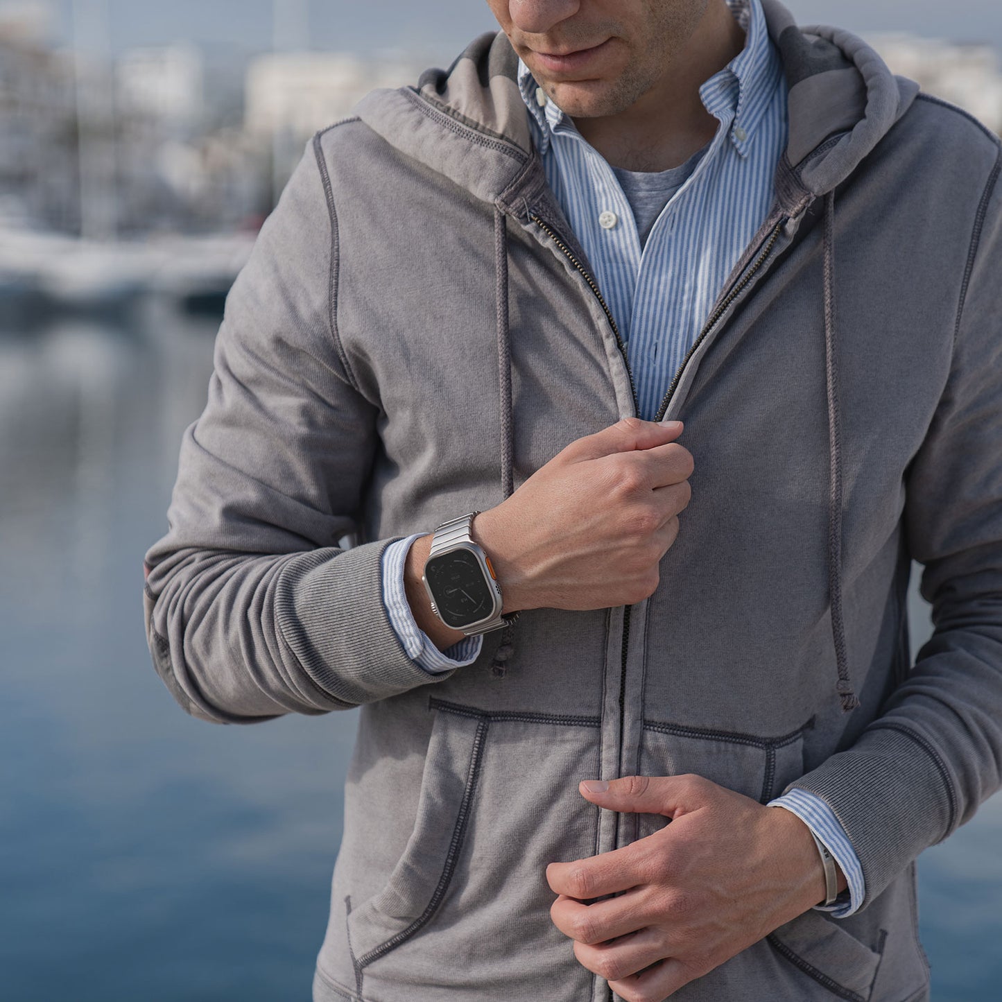 Ein Mann in einem grauen Kapuzenpullover und einem gestreiften Hemd steht am Wasser und trägt ein arktisband Apple Watch Ultra Edelstahl Armband „Commander“ am Handgelenk.