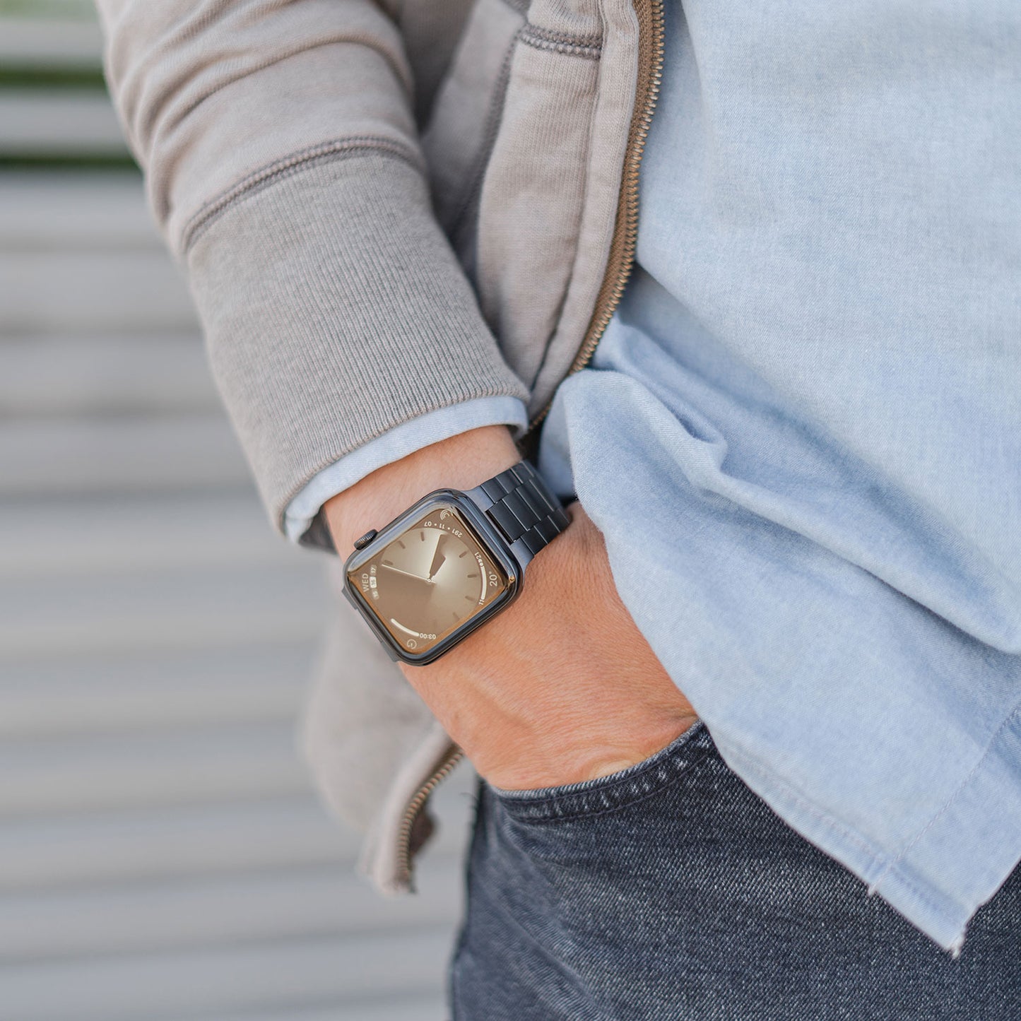 Eine Person in einem hellblauen Hemd, einem grauen Kapuzenpullover mit Reißverschluss und dunklen Jeans steht mit einer Hand in der Tasche da und zeigt ein Arktisband Apple Watch Edelstahl Gliederarmband „Edition S“ mit schlichtem, minimalistischem Design.