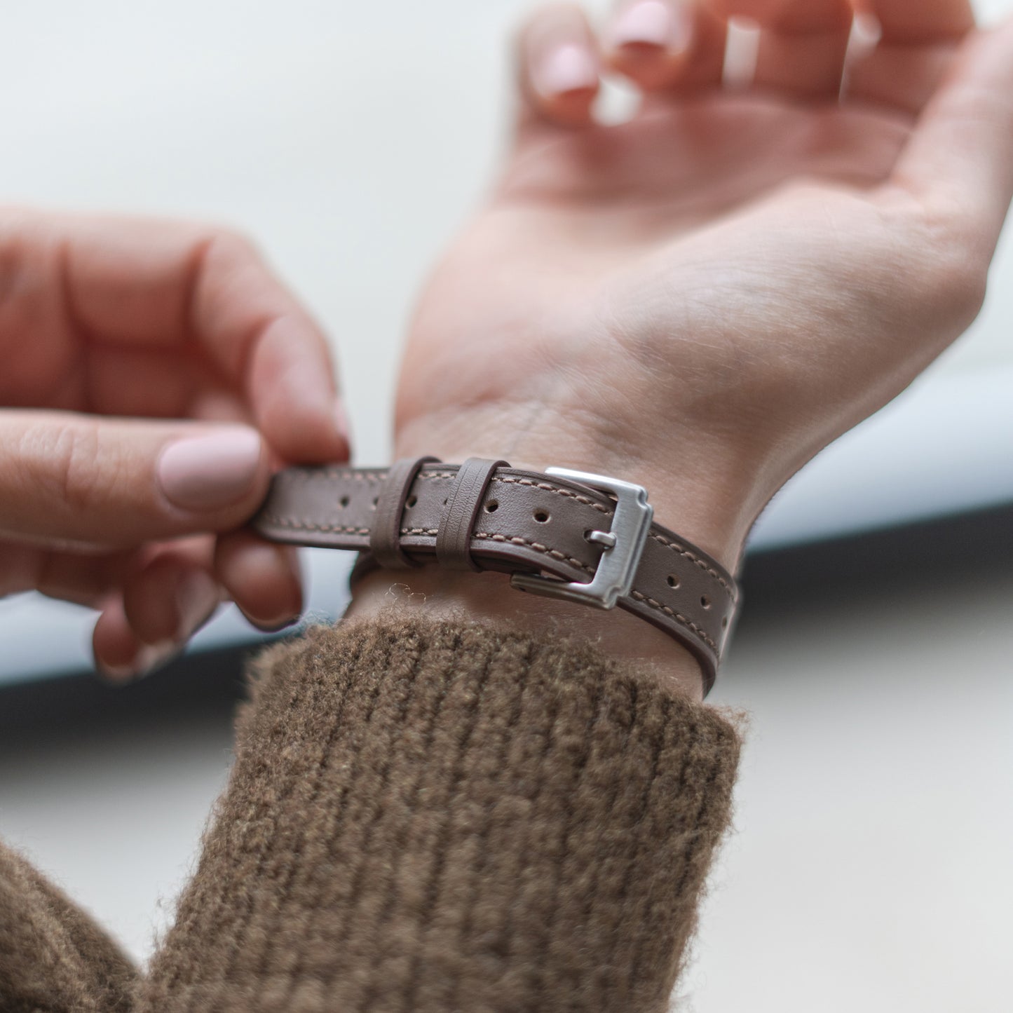 Eine Person befestigt ein Arktisband Apple Watch Lederarmband „Napoli“ am Handgelenk, trägt einen braunen Strickpullover und hat neutral lackierte Nägel.