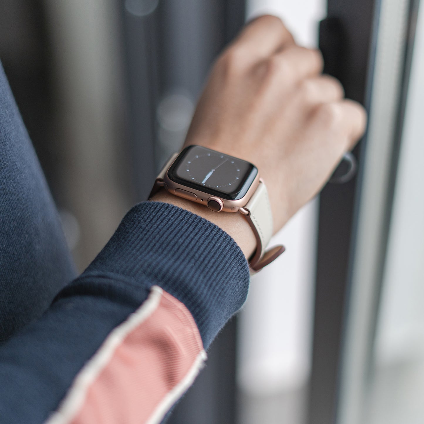 Jemand trägt das arktisband Apple Watch Lederarmband „Napoli“ von arktisband mit beigem Band und roségoldenem Gehäuse und öffnet eine Glastür, gekleidet in einen blauen Pullover mit rosa und weißen Streifen für einen minimalistischen Stil.