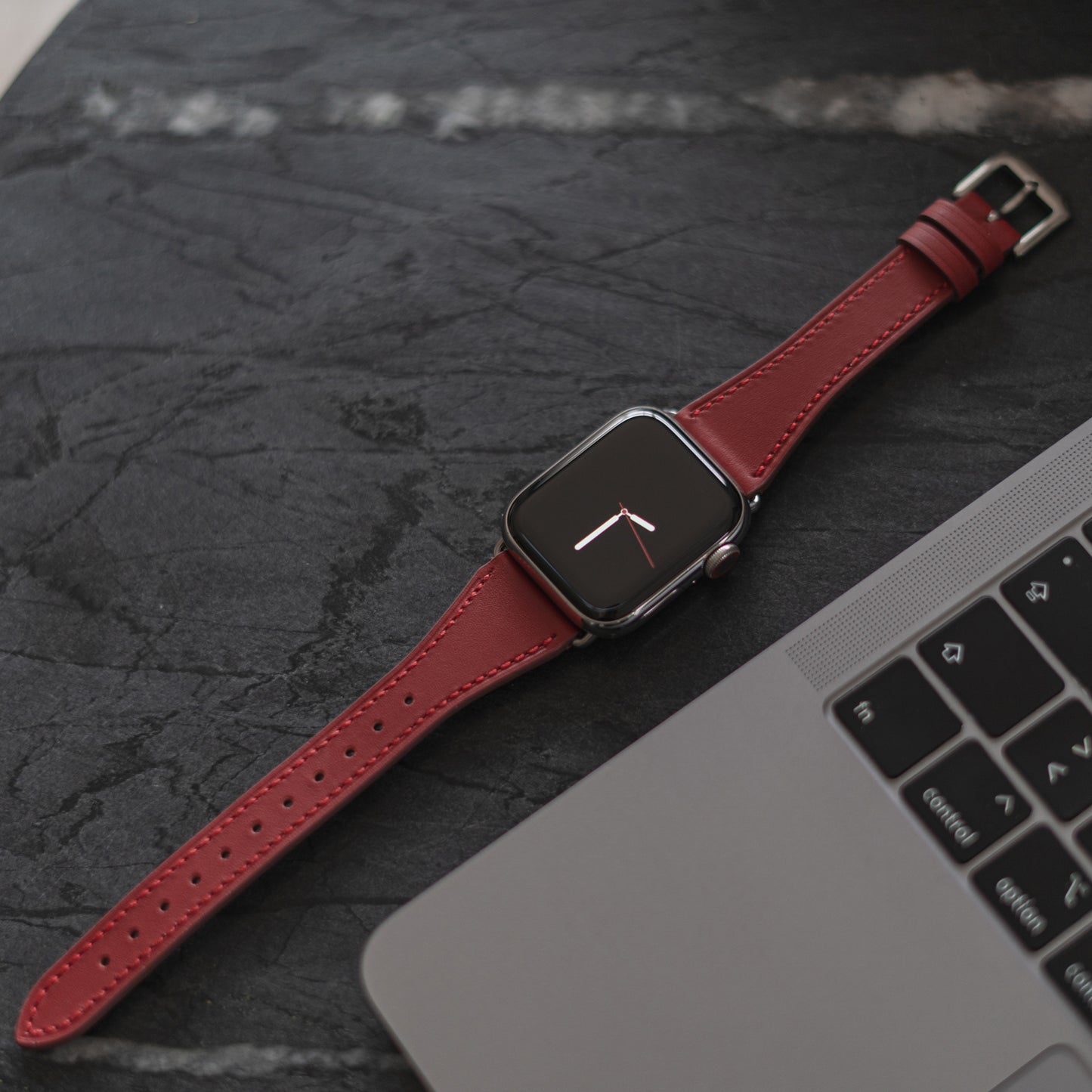 Eine Apple Watch mit dem Arktisband Apple Watch Lederarmband „Napoli“ in Rot liegt auf einer dunklen Marmoroberfläche neben einer offenen Laptoptastatur und zeigt ein einfaches analoges Zifferblatt für einen minimalistischen Stil.