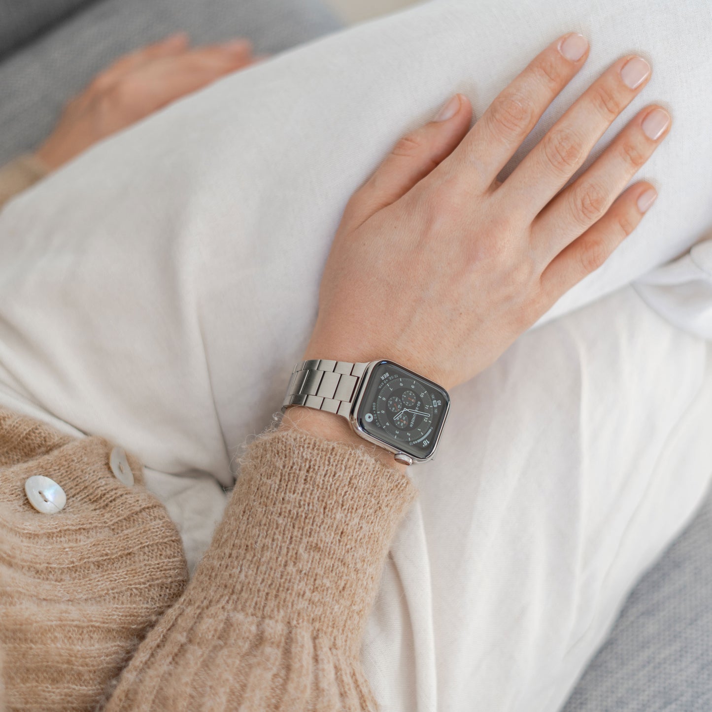 Eine Person, die das arktisband Apple Watch Edelstahl Gliederarmband „Edition S“ von arktisband mit digitalem Display trägt, ruht mit der Hand auf einer weißen Hose und sitzt in einem beigen Strickpullover mit minimalistischem Design auf einem Sofa.
