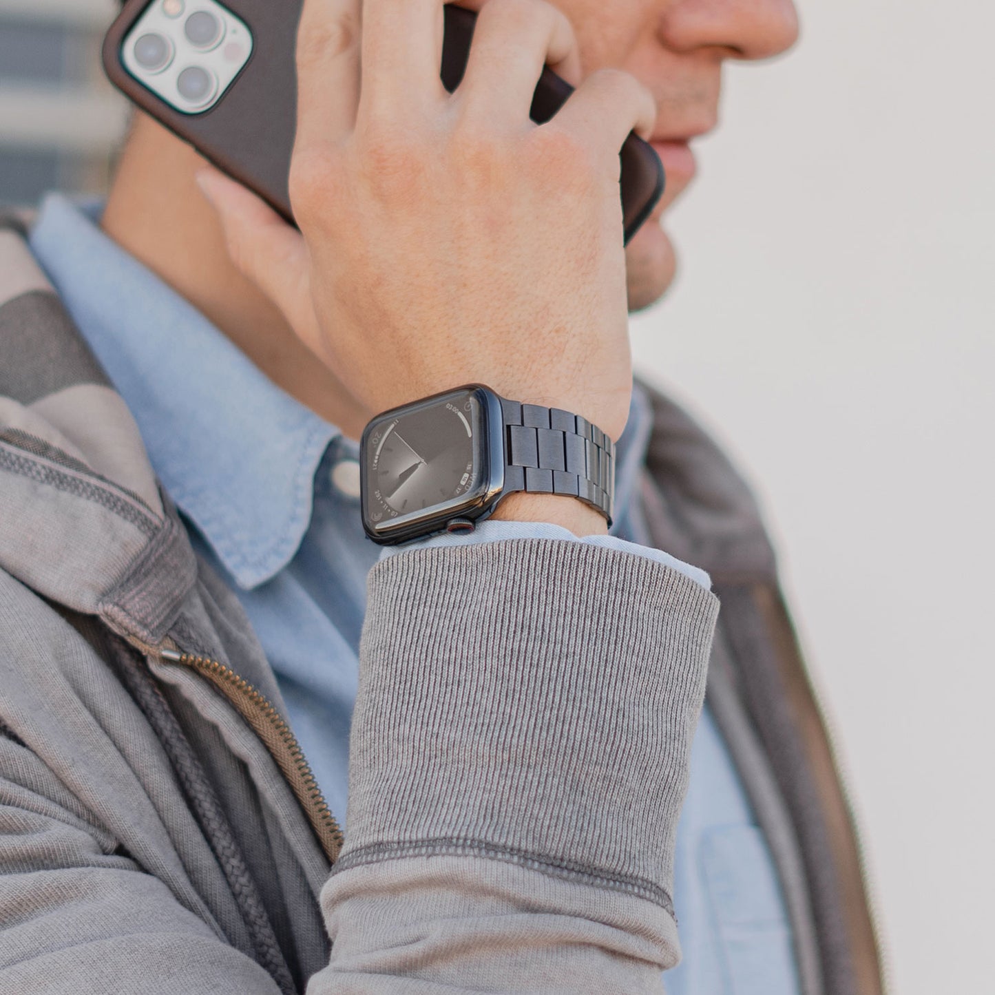 Eine Person in grauer Jacke und blauem Hemd hält ein Smartphone ans Ohr. Am Handgelenk trägt sie ein Arktisband Apple Watch Edelstahl Gliederarmband „Edition S“ und verleiht ihm einen minimalistischen Stil. Das Zifferblatt ist teilweise aus dem Bild entfernt.