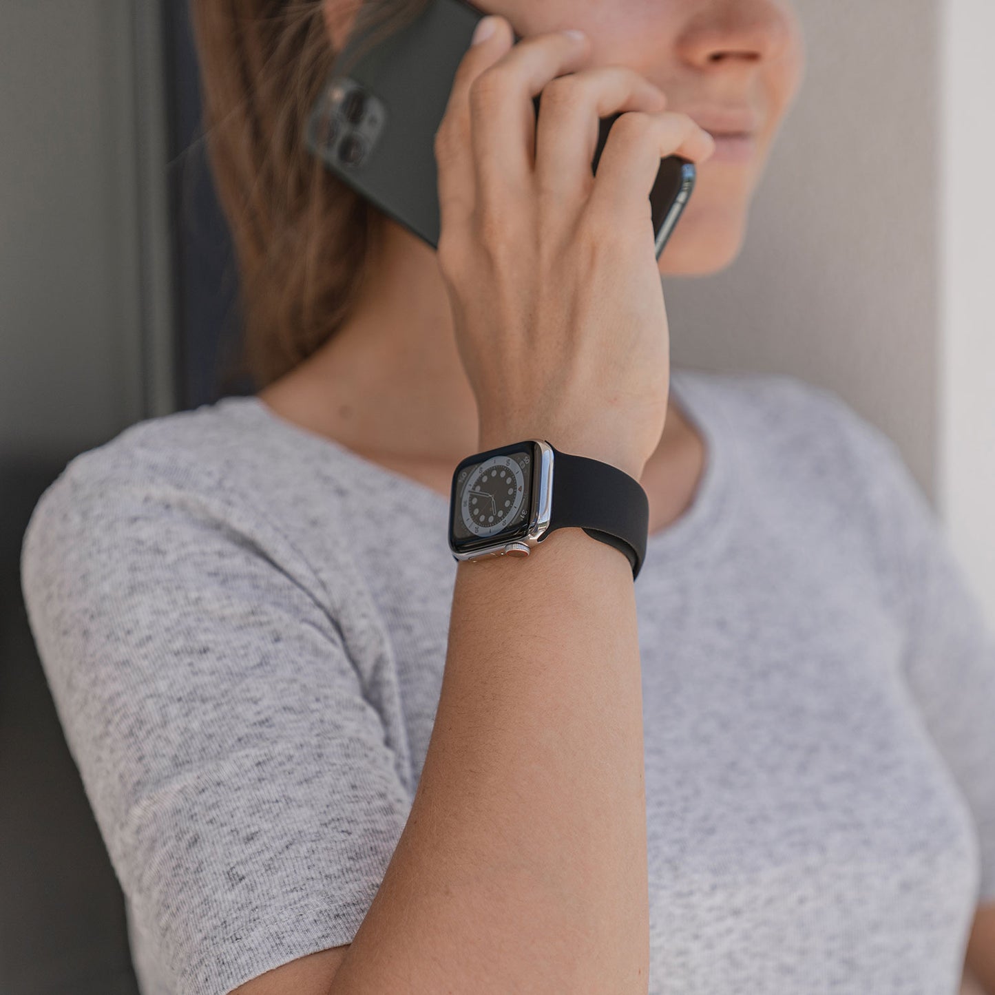 Eine Person in einem grauen T-Shirt hält ein Smartphone ans Ohr und trägt das arktisband Apple Watch Silikonarmband „Aqua“ von arktisband am Handgelenk.