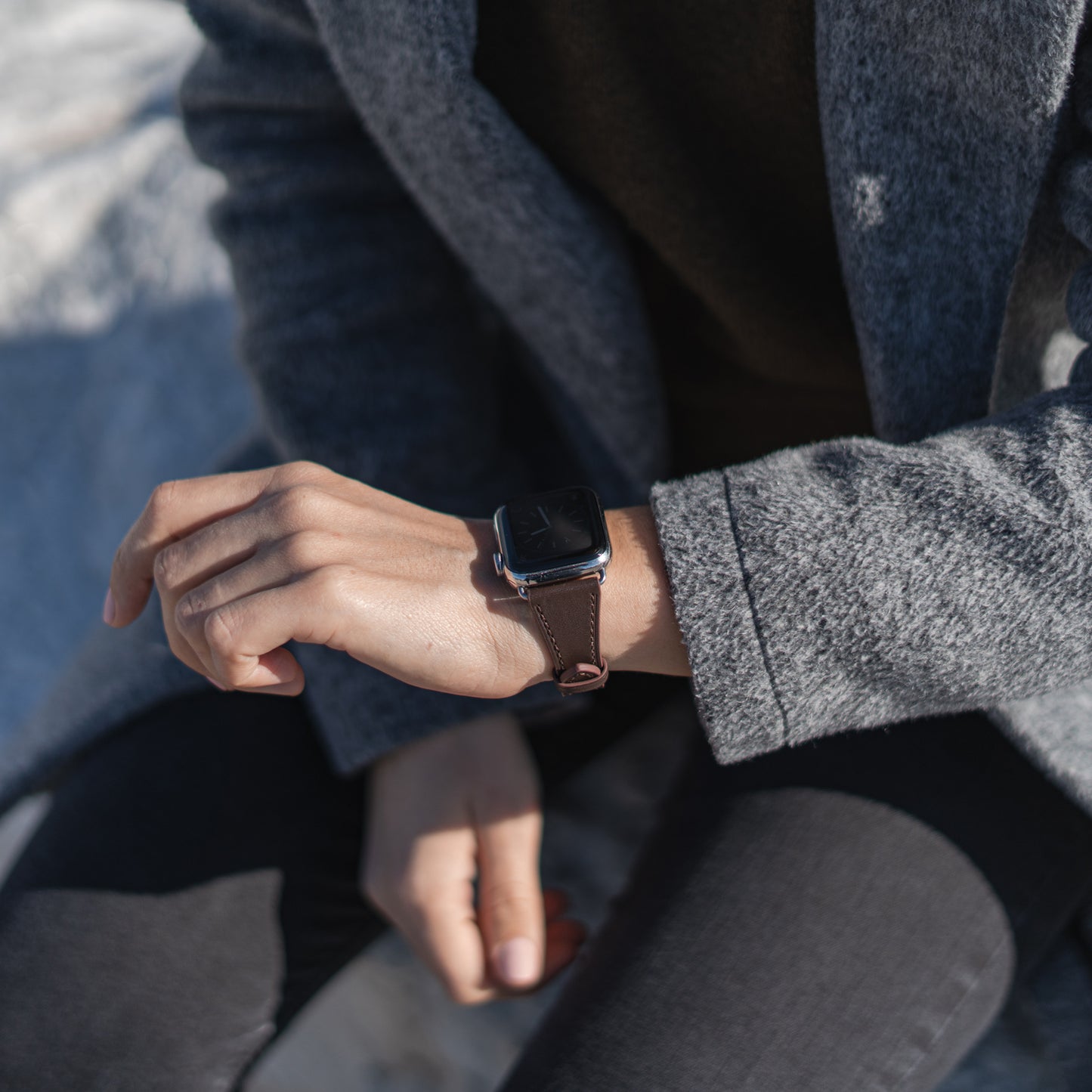 Eine Person in grauem Mantel und dunkler Hose sitzt im Freien, den linken Arm auf dem Bein, und trägt das arktisband Apple Watch Lederarmband „Napoli“ von arktisband aus echtem Leder mit einer eleganten Edelstahl-Dornschließe.