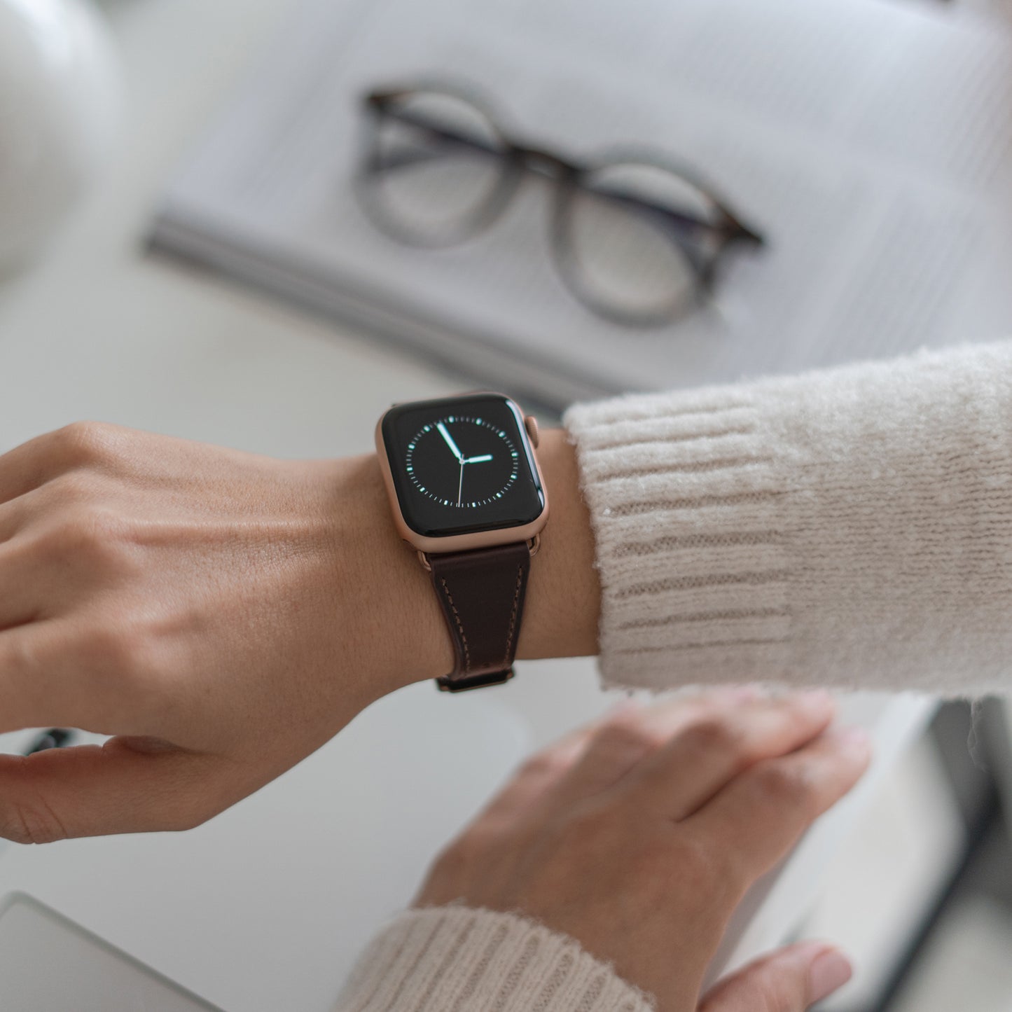 Eine Person trägt eine minimalistische Smartwatch mit dem Arktisband Apple Watch Lederarmband „Napoli“ in Braun und schaut auf die Uhrzeit. Sie trägt einen leichten Pullover und sitzt an einem Schreibtisch. Im Hintergrund sieht man eine Brille und ein offenes Buch.