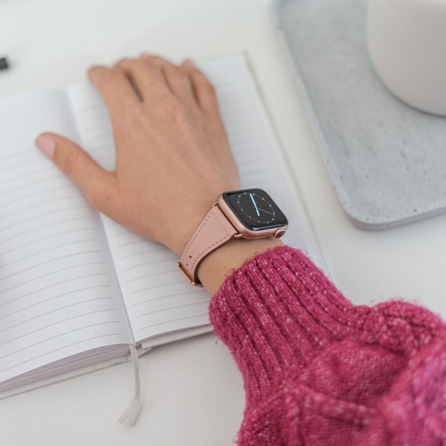 Eine Person mit dem Arktisband Apple Watch Lederarmband „Napoli“ schreibt in ein offenes liniertes Notizbuch und trägt einen rosa Strickpullover. Ein graues Tablett im Hintergrund unterstreicht den minimalistischen Stil.