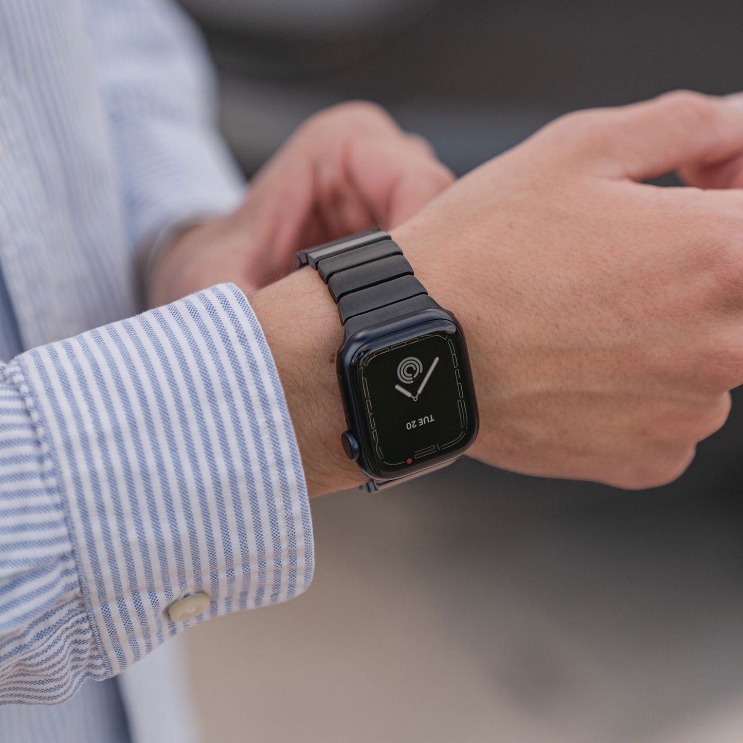 Eine Person in einem blau-weiß gestreiften Hemd richtet ein arktisband Apple Watch Edelstahl Gliederarmband „Iconic“ von arktisband; auf dem Display der Uhr werden die Uhrzeit und eine Standort-PIN angezeigt, was auf die Kompatibilität mit der Apple Watch hinweist.