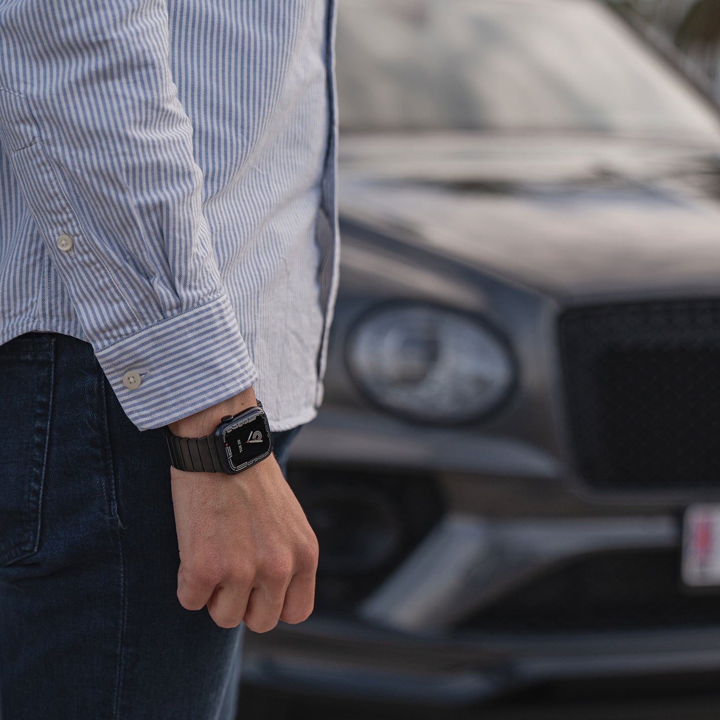 Eine Person in blau gestreiftem Hemd und Jeans, die das Arktisband Apple Watch Edelstahl Gliederarmband „Iconic“ trägt, steht vor einem verschwommenen grauen Luxusauto. Nur ihr Arm und ein Teil ihres Oberkörpers sind zu sehen.