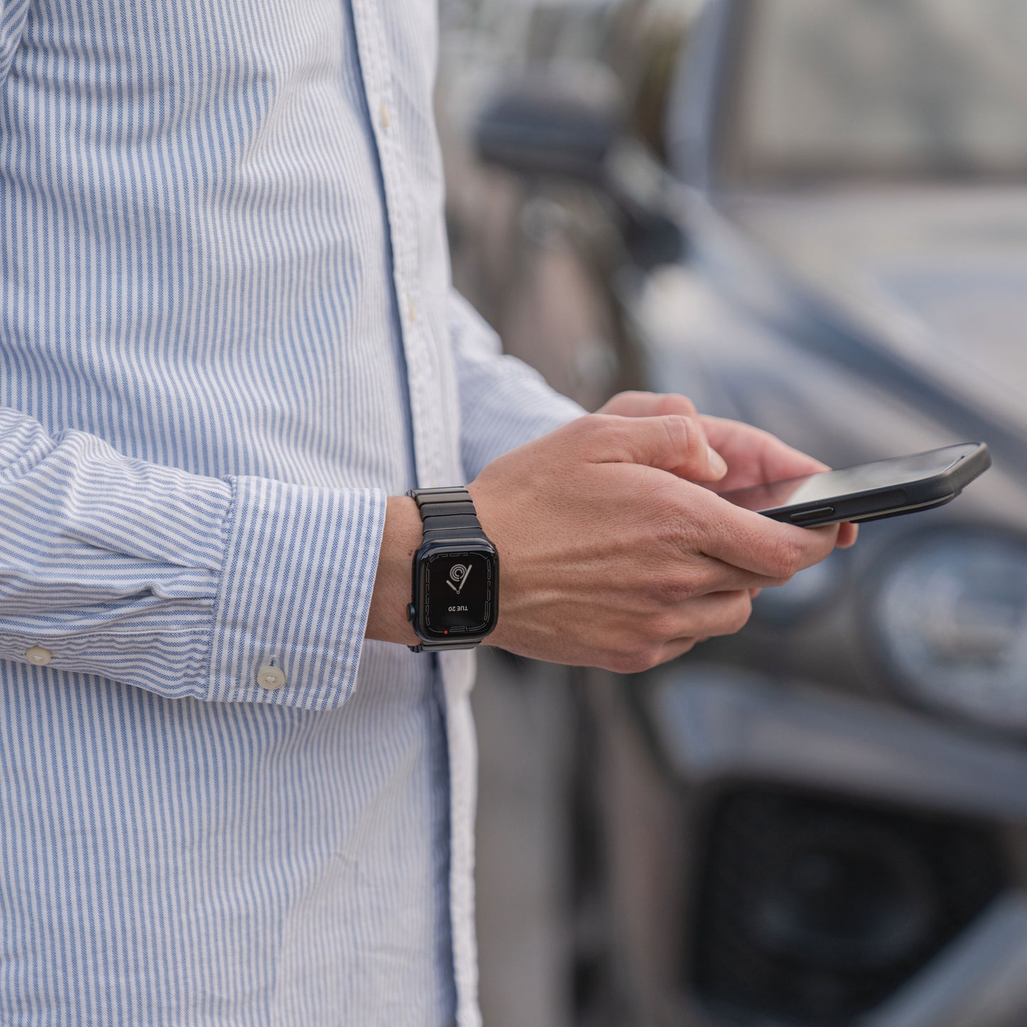 Eine Person in einem gestreiften Hemd nutzt im Freien ein Smartphone. Am linken Handgelenk trägt sie das Arktisband Apple Watch Edelstahl Gliederarmband „Iconic“. Im unscharfen Hintergrund ist ein Auto zu sehen.