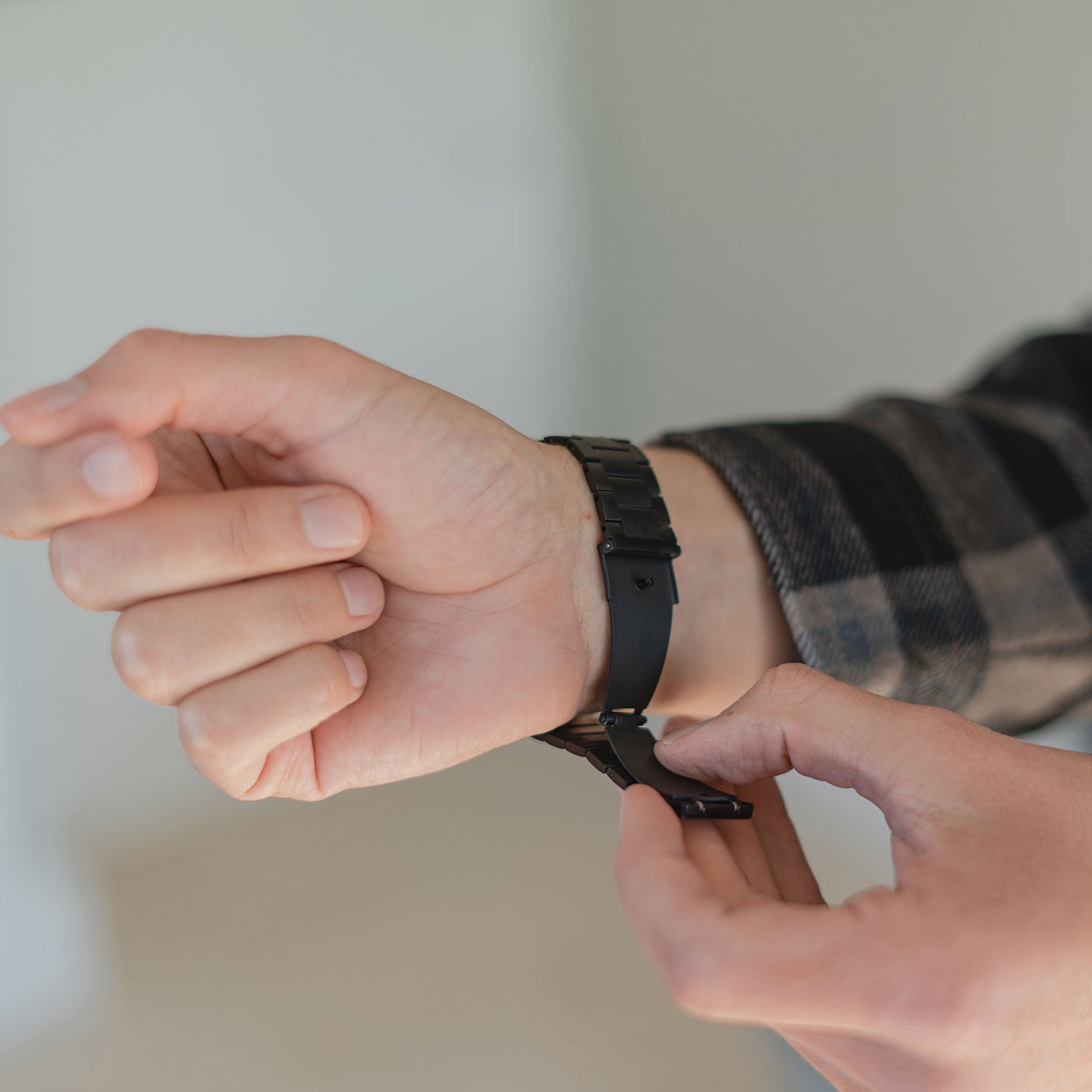 Eine Person im karierten Hemd schließt vor unscharfem Hintergrund den Verschluss eines Arktisband Apple Watch Edelstahl Gliederarmbands „Edition S“ um ihr linkes Handgelenk.