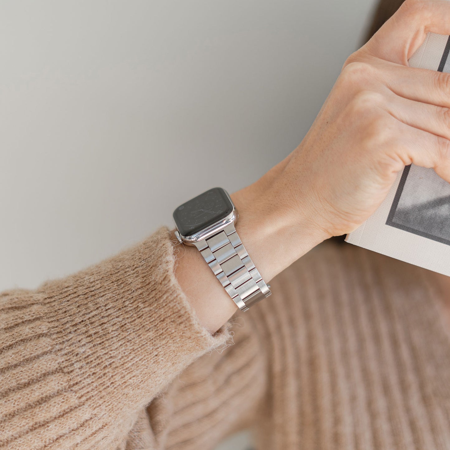 Eine Person trägt das arktisband Apple Watch Edelstahl Gliederarmband „Edition S“ mit minimalistischem Design und hält ein Buch. Ein beiger Strickpullover betont das elegante Armband, der Fokus liegt auf Handgelenk und Hand.