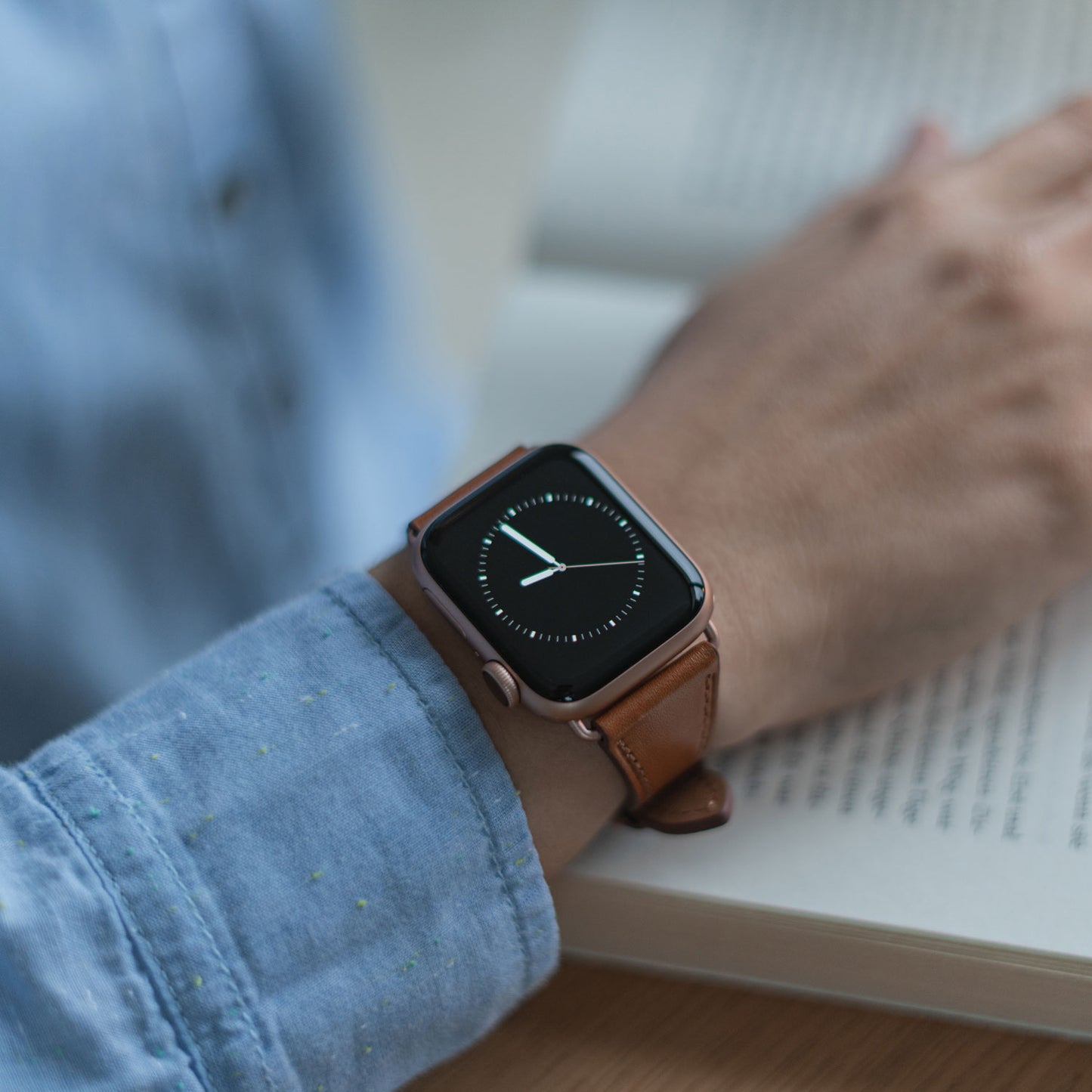 Eine Person, die das arktisband Apple Watch Lederarmband „Napoli“ trägt, legt ihren Arm auf ein offenes Buch. Ihr hellblaues, gesprenkeltes Langarmshirt unterstreicht einen minimalistischen Stil.