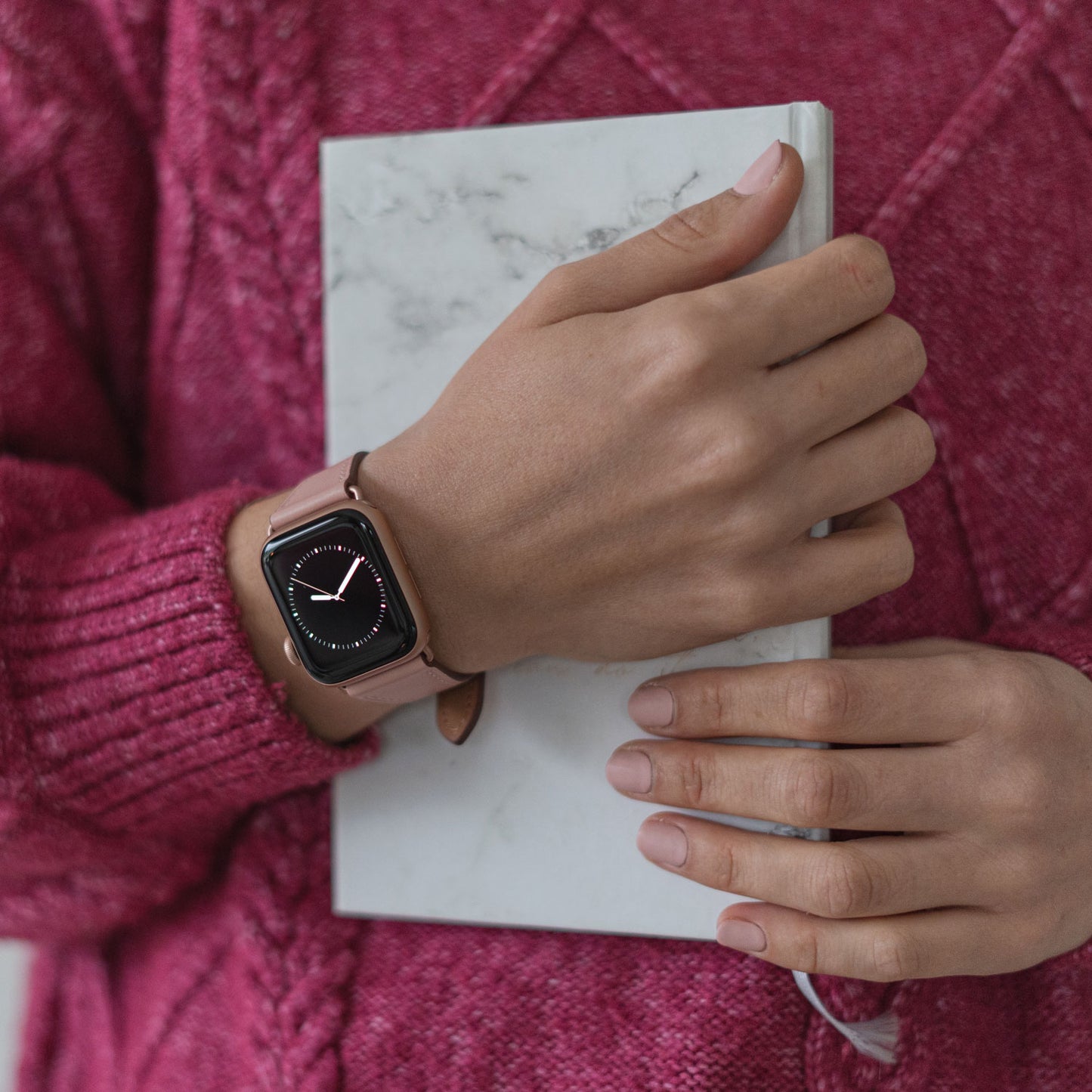 Eine Person, die das Arktisband Apple Watch Lederarmband „Napoli“ in Pink und einen pinkfarbenen Pullover trägt, hält ein geschlossenes Notizbuch mit Marmormuster in der Hand, ihre hellpinken Nägel betonen den minimalistischen Stil.
