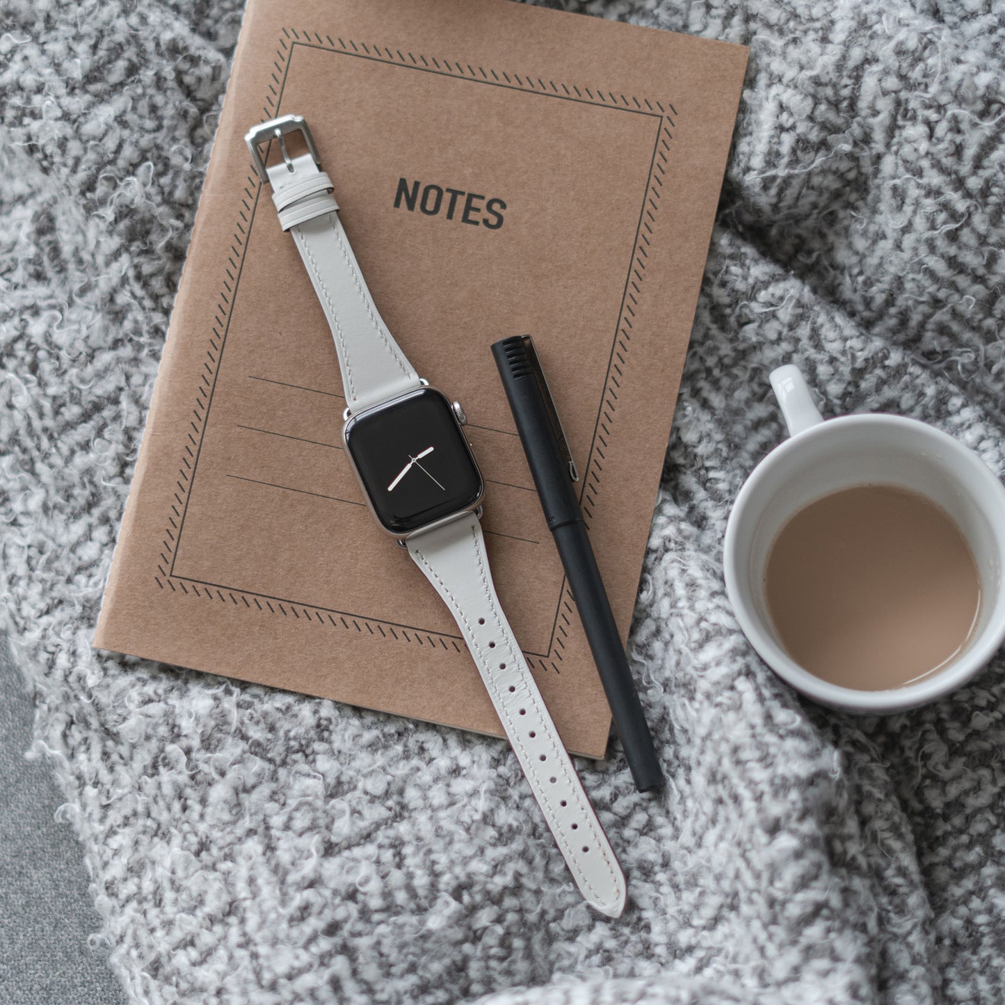 Ein geschlossenes braunes Notizbuch mit der Aufschrift NOTES, ein schwarzer Stift, ein Arktisband Apple Watch Lederarmband „Napoli“ und eine weiße Tasse mit hellem Kaffee ruhen auf einer grauen strukturierten Decke für einen minimalistischeren Stil.