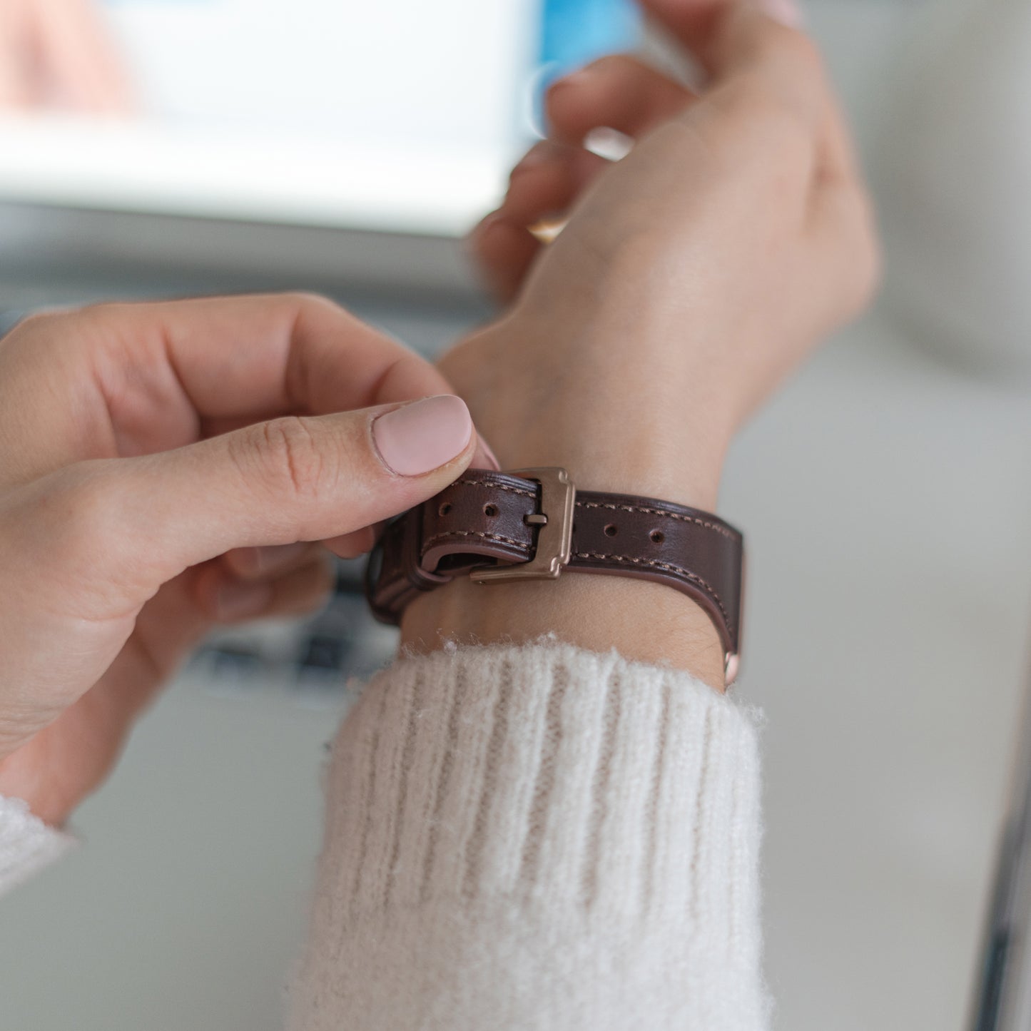 Eine Person im weißen Pullover befestigt das arktisband Apple Watch Lederarmband „Napoli“ um ihr Handgelenk und zeigt sich dabei im minimalistischen Stil; im Hintergrund sind ein unscharfer Laptop und andere Gegenstände zu sehen.