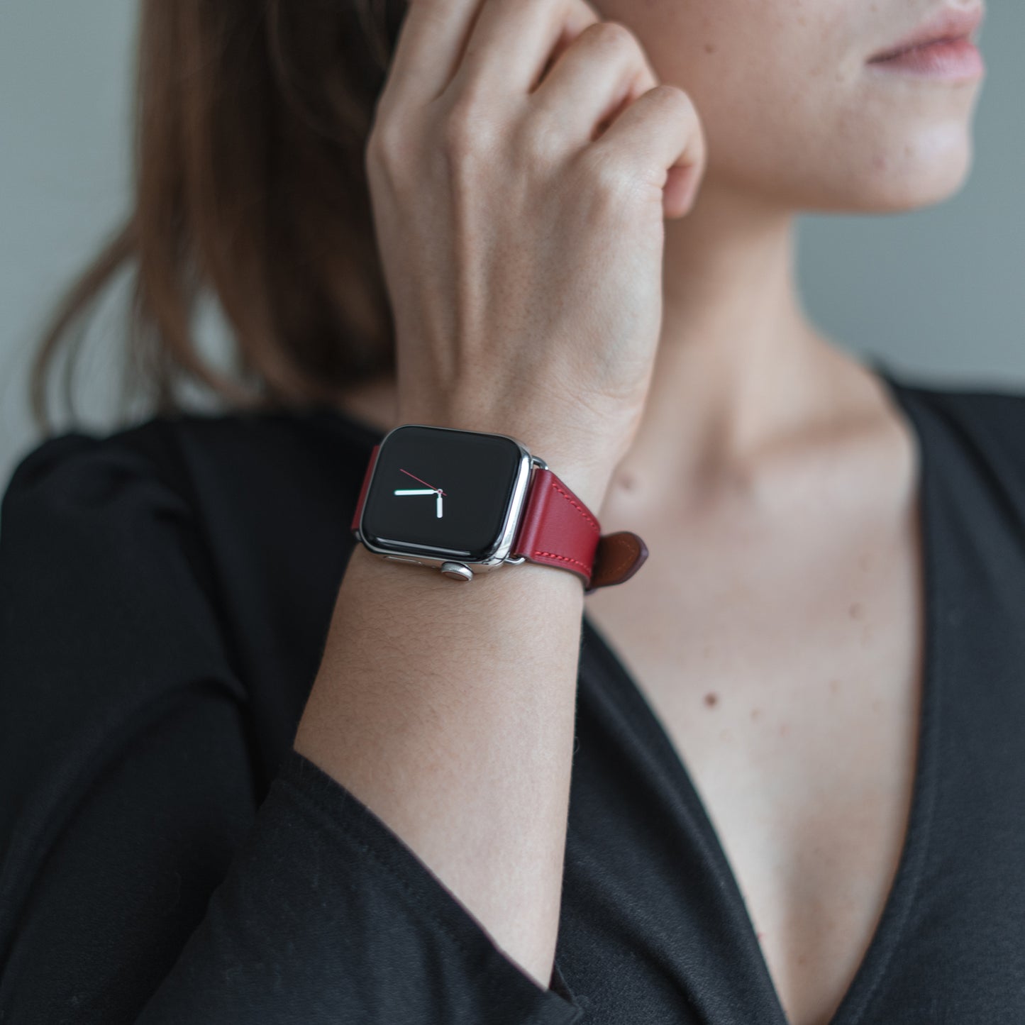 Eine Person trägt ein schwarzes V-Ausschnitt-Top und das arktisband Apple Watch Lederarmband „Napoli“ im minimalistischen Stil mit rotem Band von arktisband. Das analoge Zifferblatt wird angezeigt, wenn sie ihr Ohr berührt, das Zifferblatt ist teilweise sichtbar.