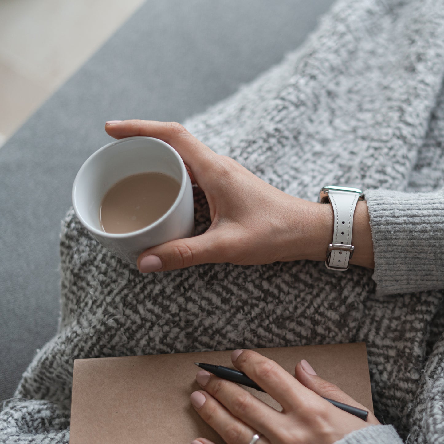 Eine Person, die einen grauen Pullover und das arktisband Apple Watch Lederarmband „Napoli“ von arktisband trägt, hält ein heißes Getränk und ein Notizbuch mit Stift auf einer strukturierten grauen Decke und verkörpert damit einen gemütlichen, minimalistischen Stil.